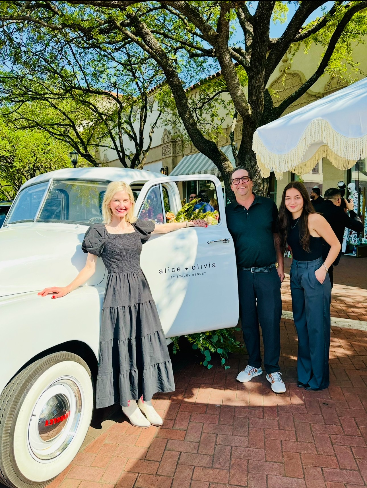Three people stand outside a vintage white car under a tree, with a white canopy in the background.