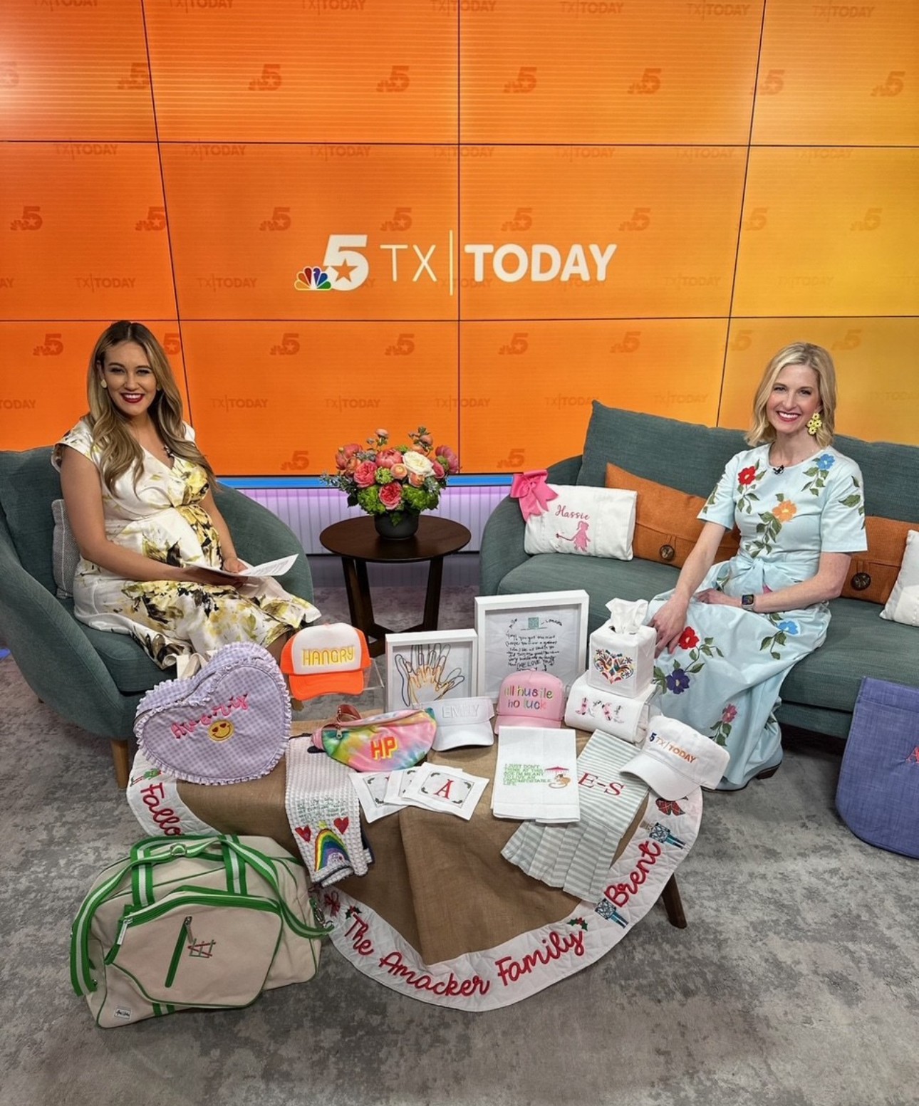 Two women sit on sofas with a table of children's drawings and a floral arrangement in front of a TV screen displaying '5 TX TODAY'.