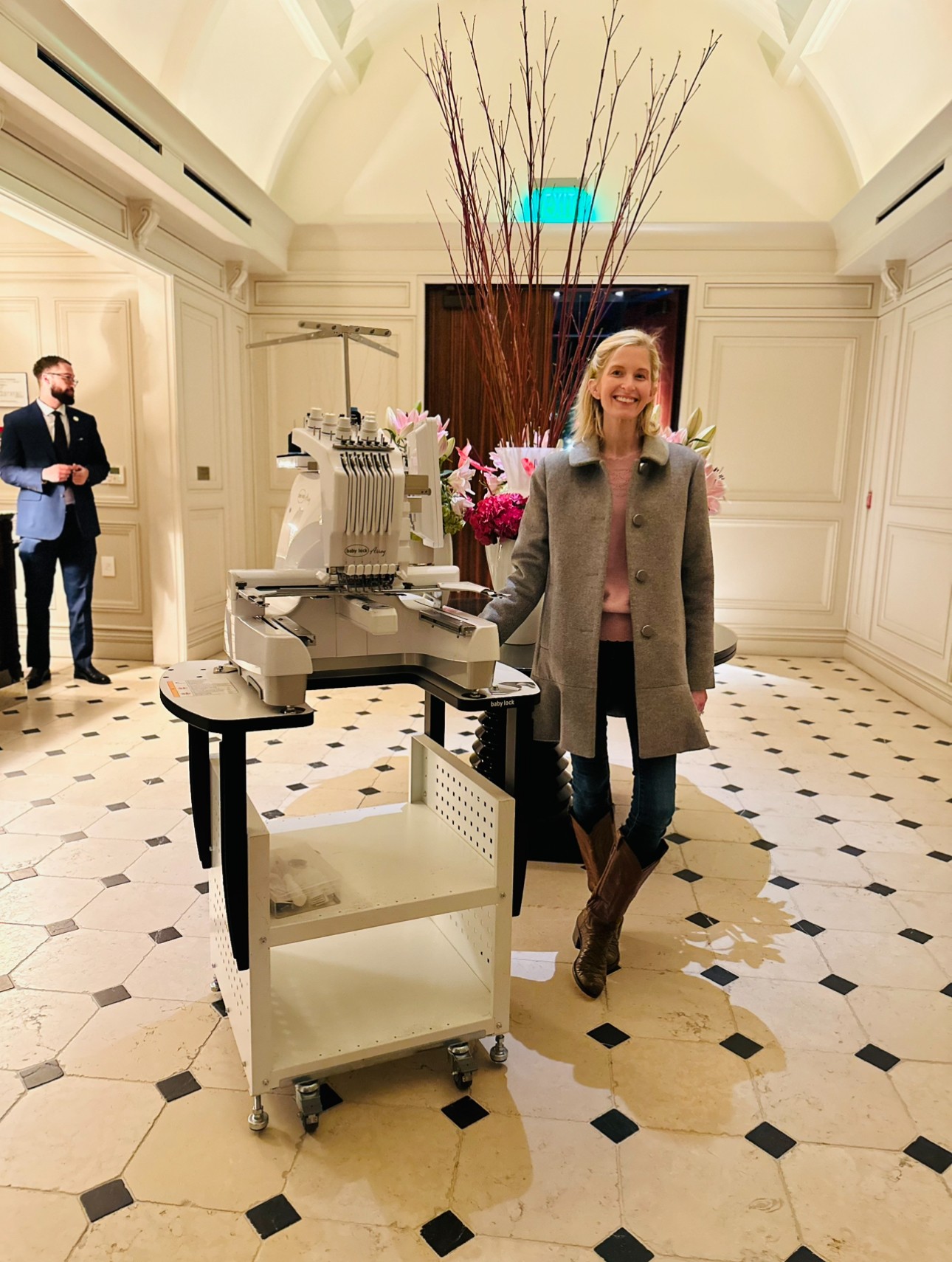 Woman standing next to a machine in a luxurious room with white walls, black and white tiled floor, and a large floral arrangement behind her.