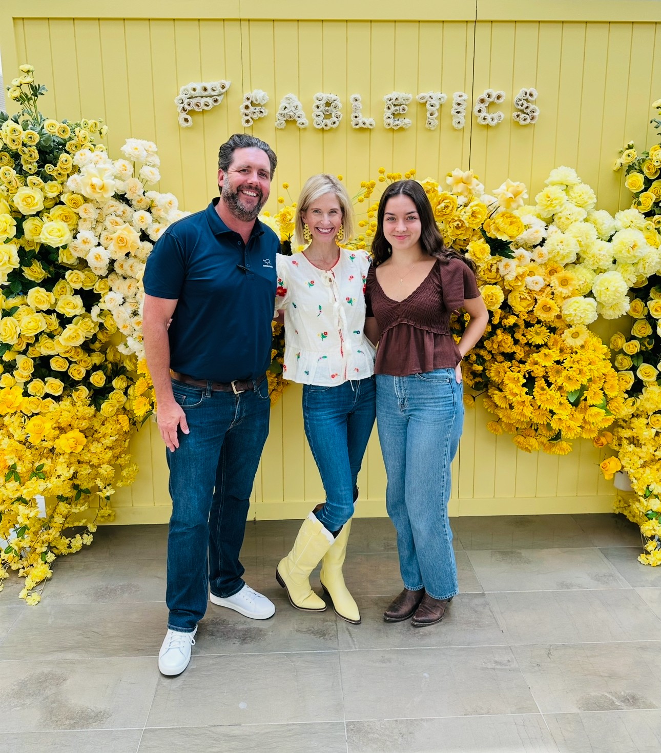 Three people standing in front of a yellow floral backdrop with the word 'GARDEN' spelled out, smiling at the camera.