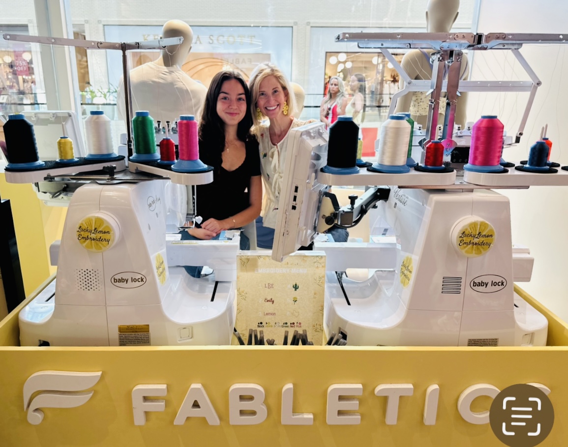Two women stand behind a sewing machine display with colorful spools of thread, smiling at the camera.