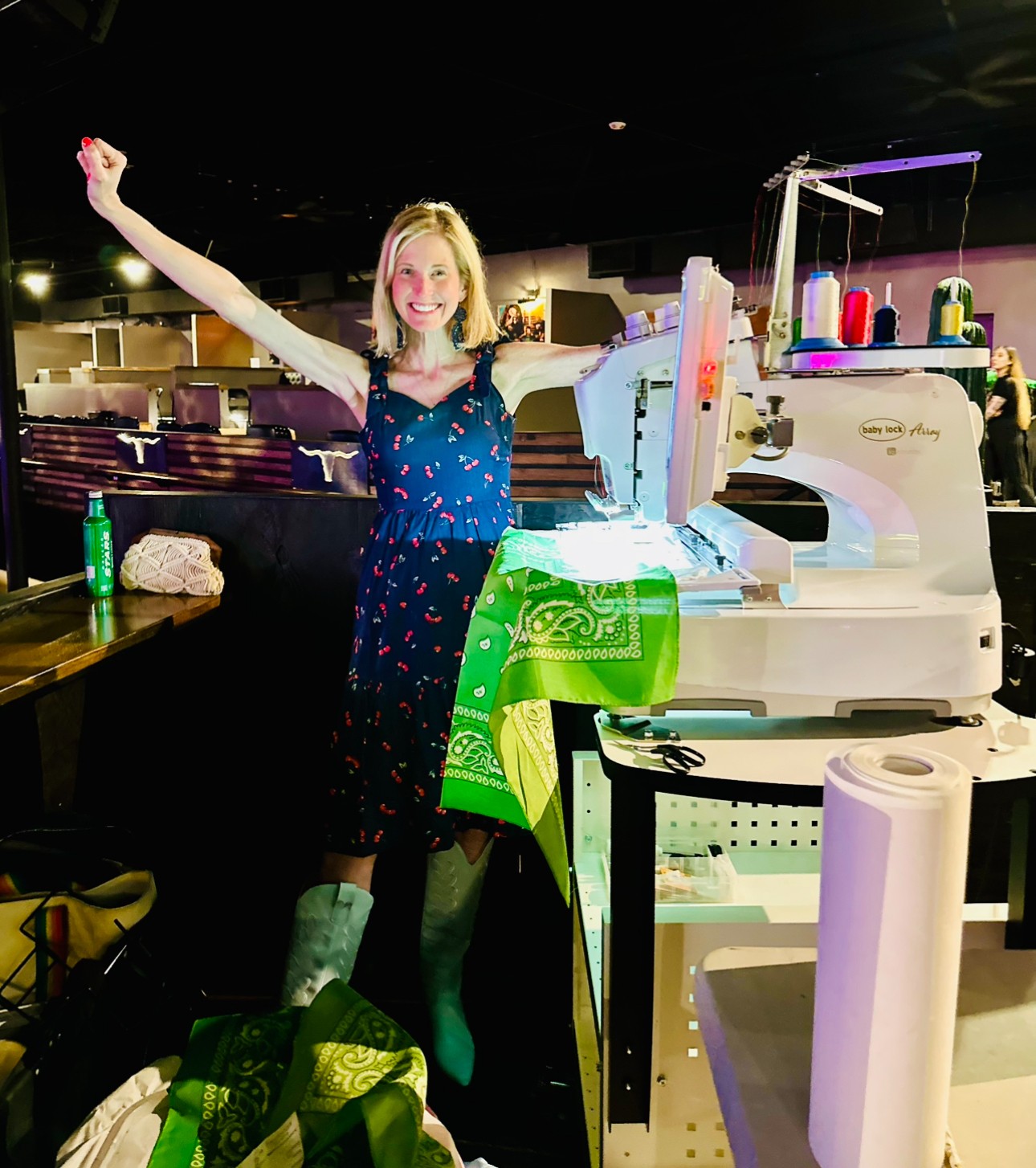 Woman smiling with arms raised, standing behind a sewing machine in a workshop, with fabric and supplies around.
