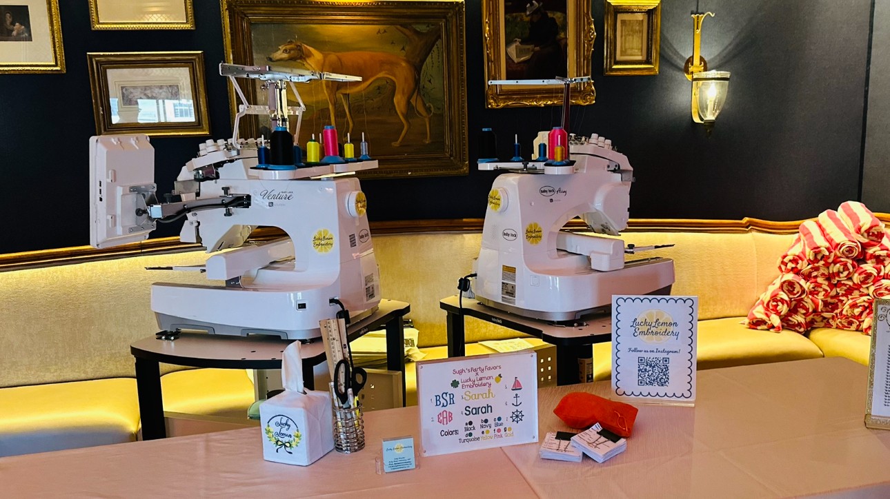 Two sewing machines on tables with various sewing supplies, in a room with framed artwork and a yellow sofa.