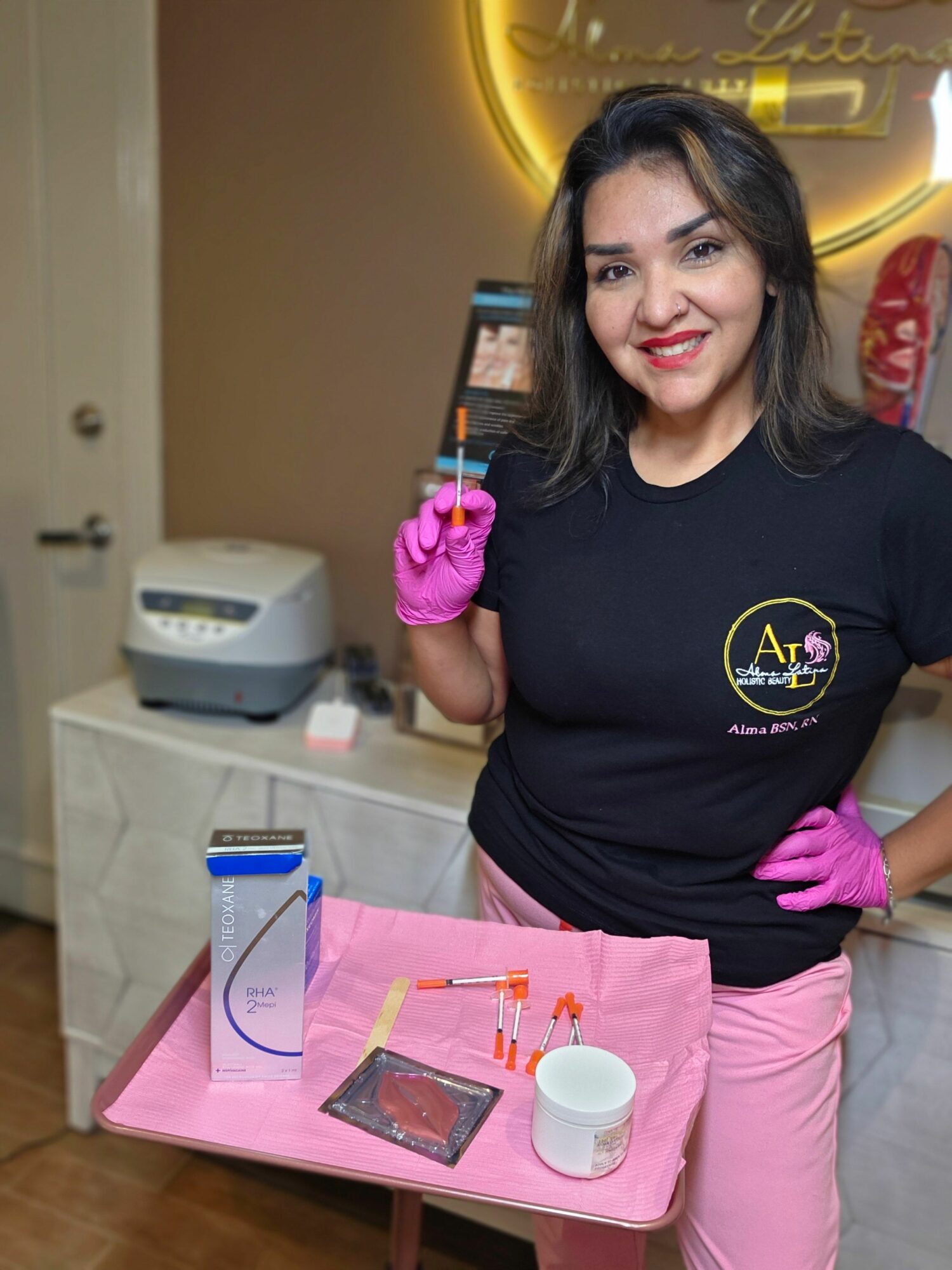 Woman with black hair wearing pink gloves and a black shirt, standing in a room with medical equipment, smiling.