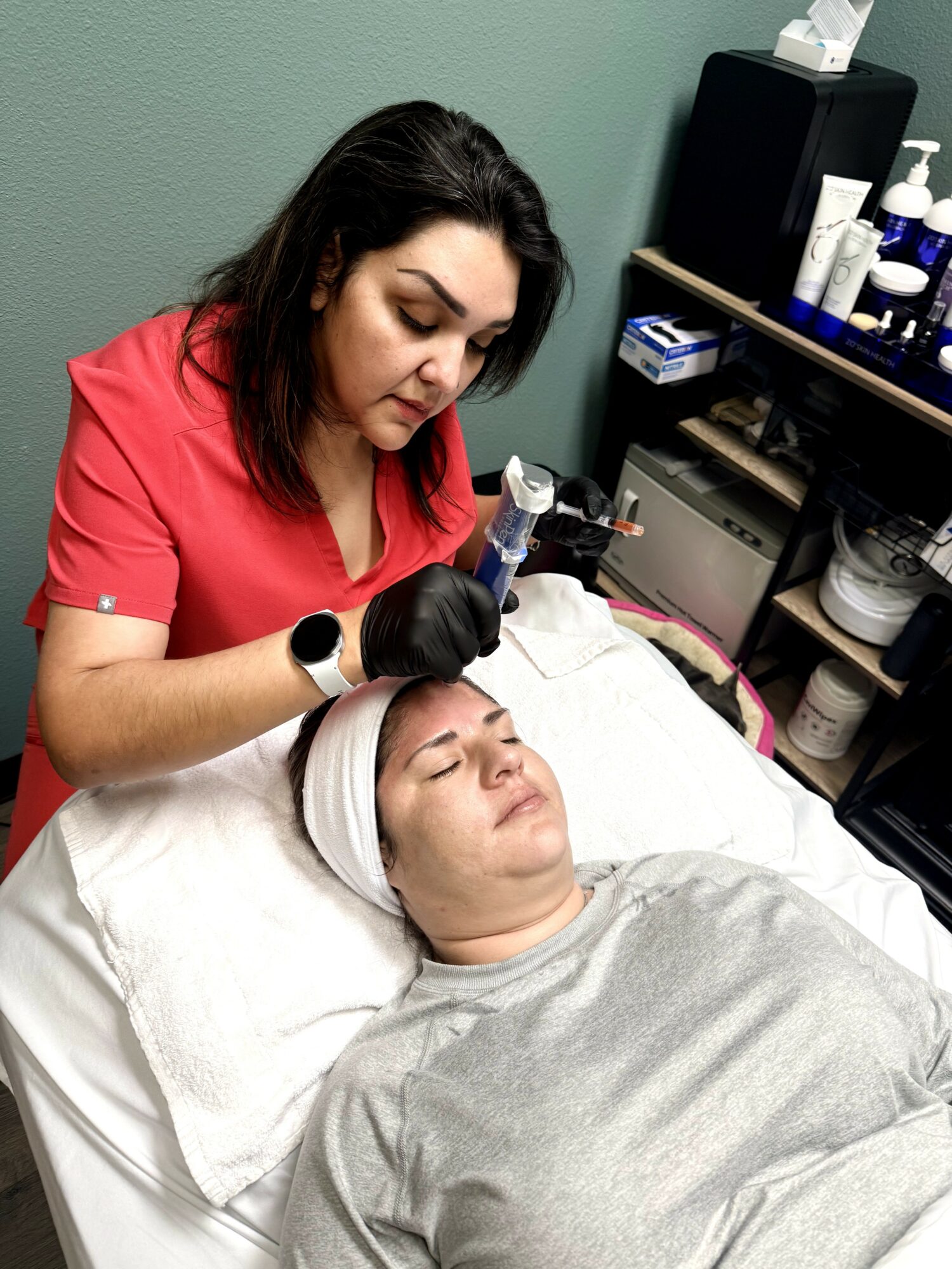 A woman receives a facial treatment from a professional in a clinic room, lying on a bed with eyes closed.
