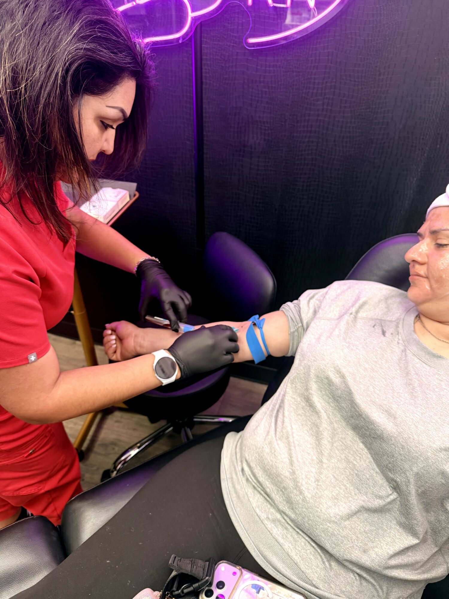 A woman in a red shirt and black gloves draws blood from a man lying in a chair, using a tourniquet on his arm.