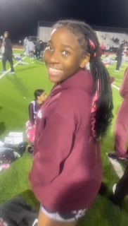 Smiling girl with braided hair in a maroon hoodie on a sports field at night.