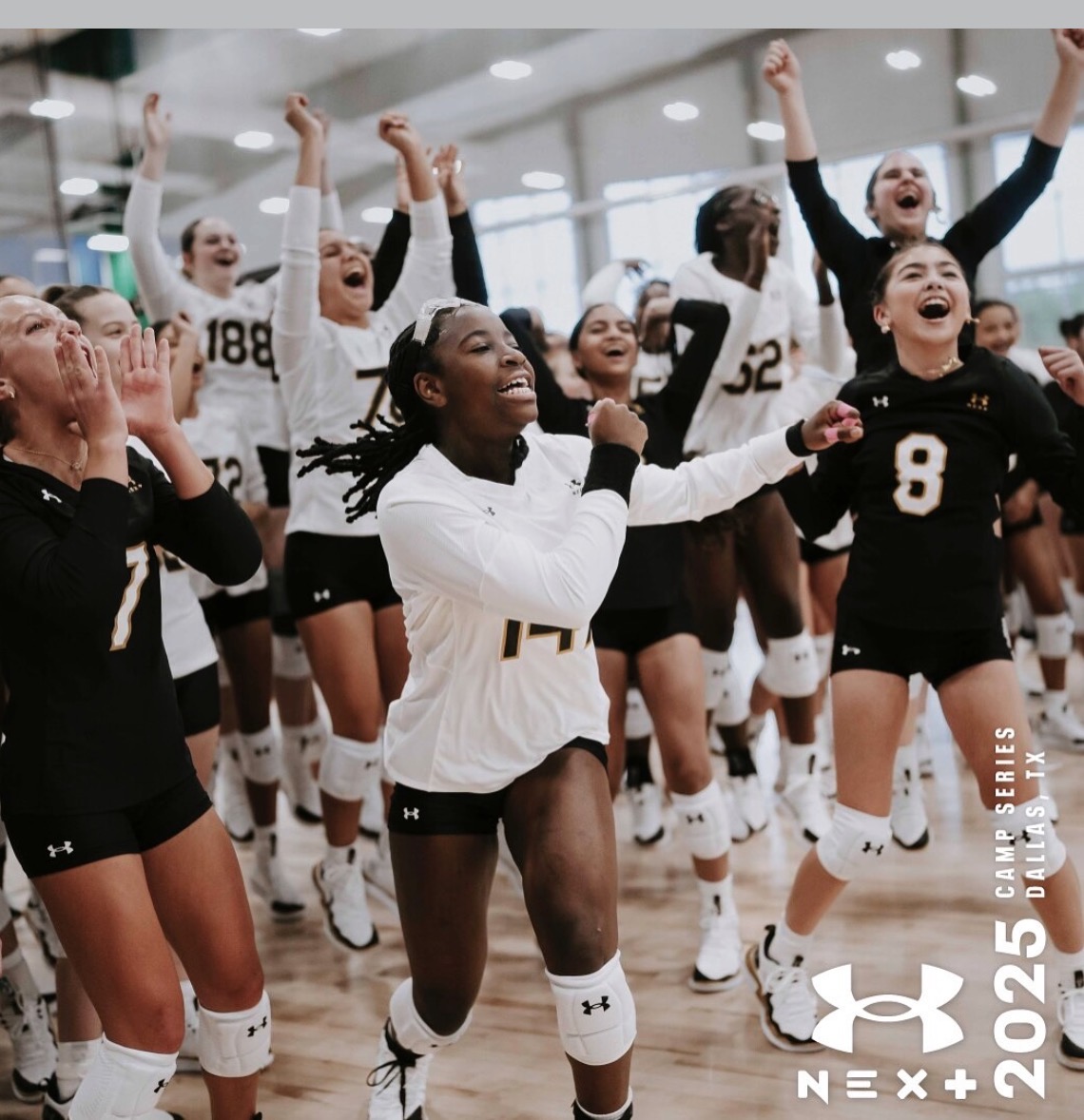Group of female volleyball players celebrating indoors, some with arms raised, smiling, wearing sports uniforms.