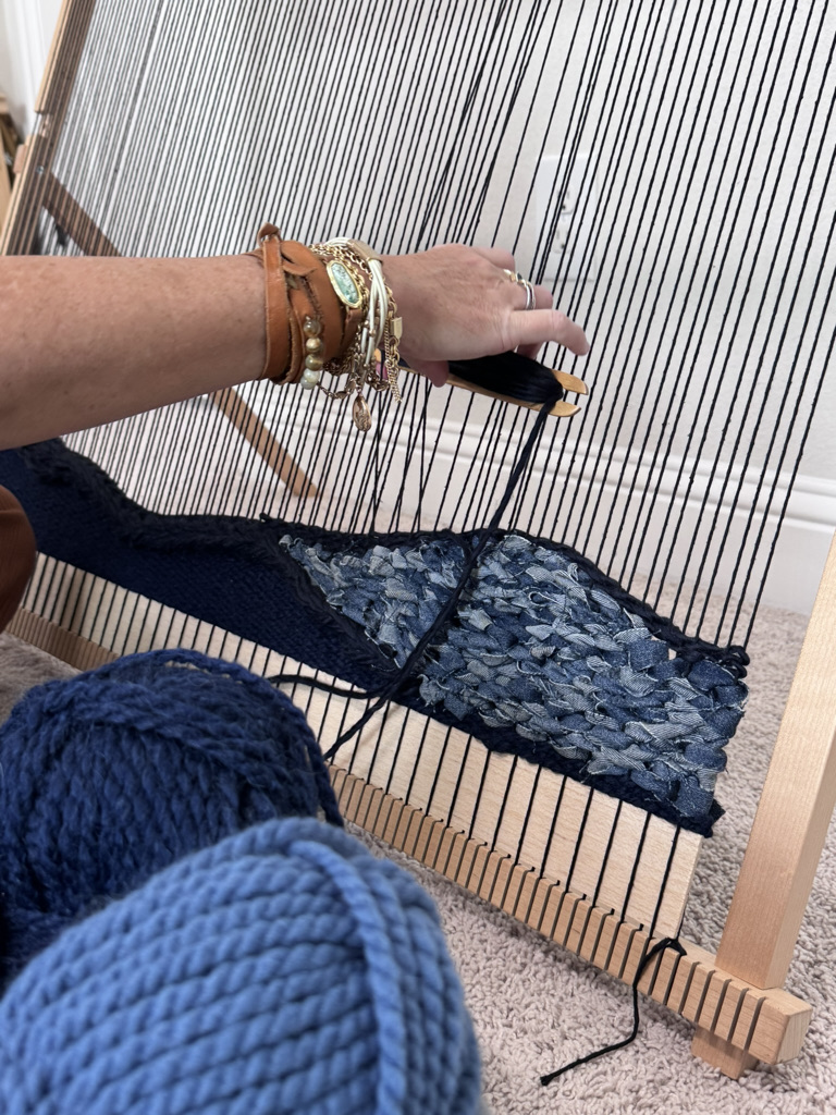 Person weaving fabric on a loom, wearing bracelets and a watch, with a blue knitted garment in the foreground.