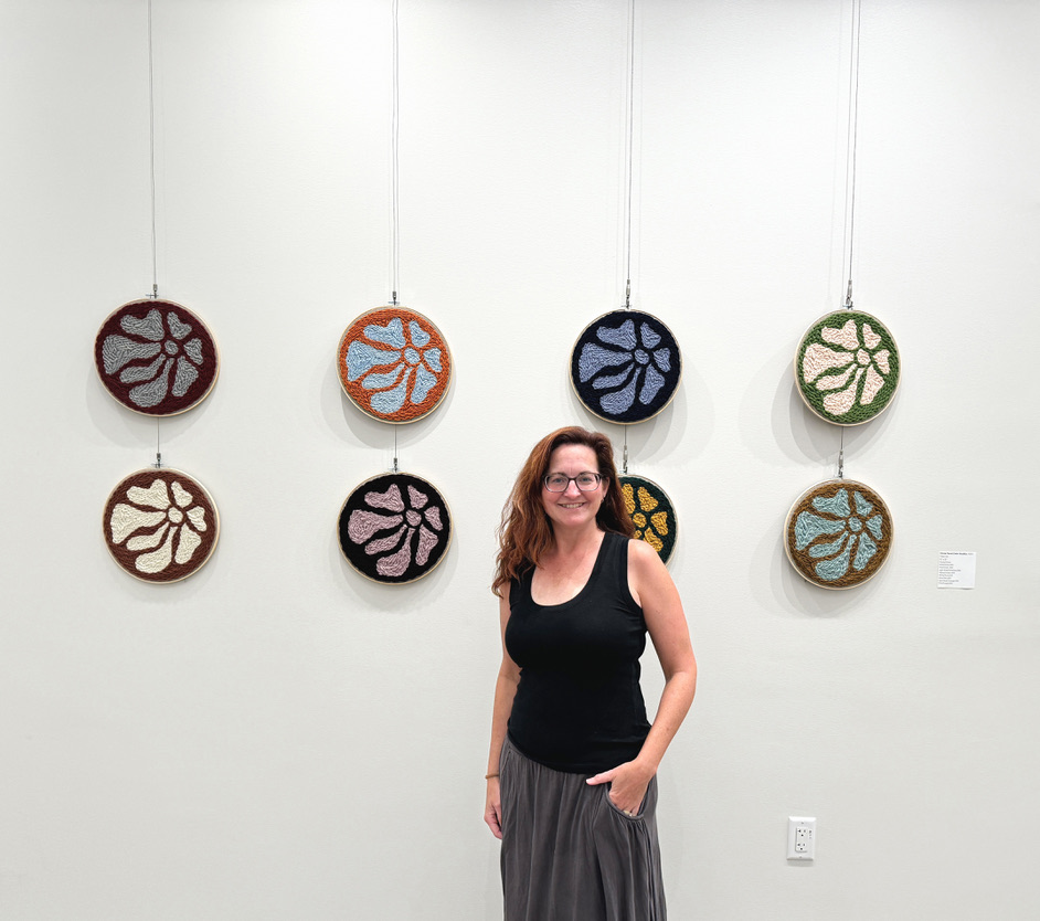 Woman standing in front of colorful circular art pieces on a white wall, smiling, with her hand in her pocket.