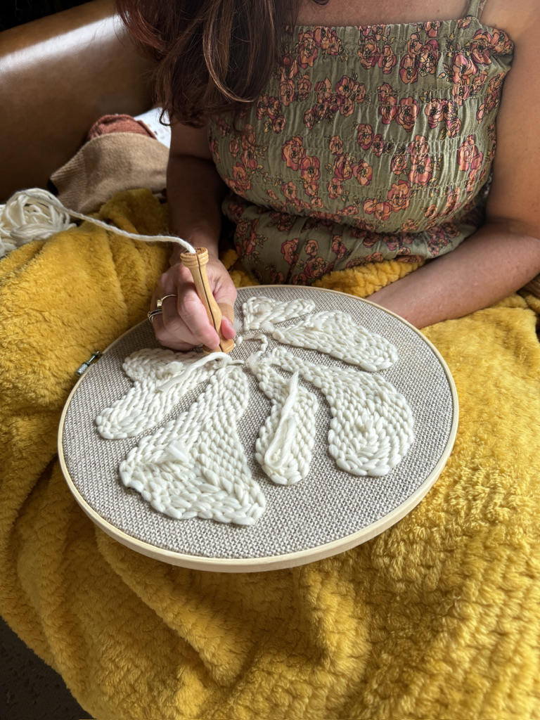 Person knitting white yarn bunny shapes on a round frame, sitting on a yellow blanket, with a patterned top visible.