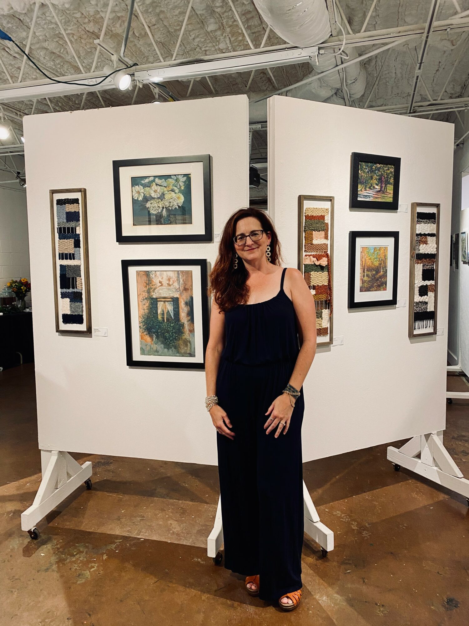 Woman in black jumpsuit standing in front of white display panels with framed artwork, in an art gallery setting.
