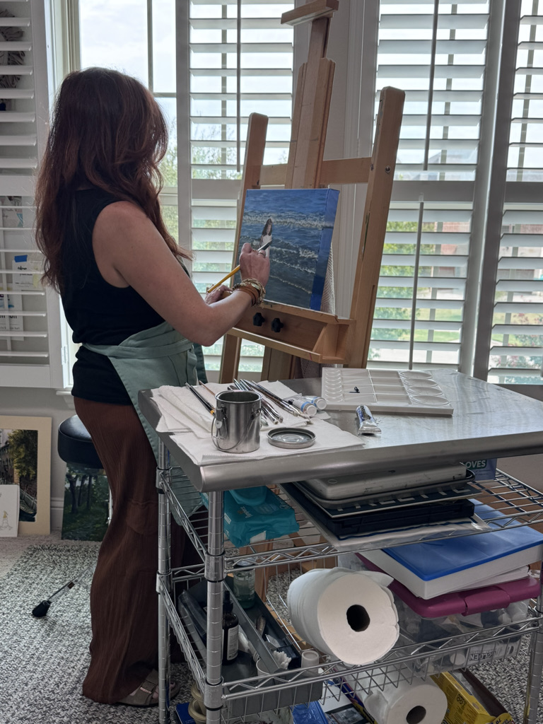 Woman painting on canvas in bright room with large windows, art supplies on table, easel, and view outside.