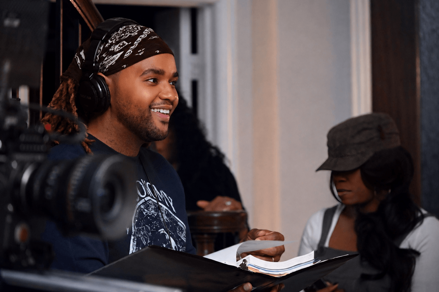 A smiling man with headphones and a bandana, holding a script, in a recording studio with a woman nearby.