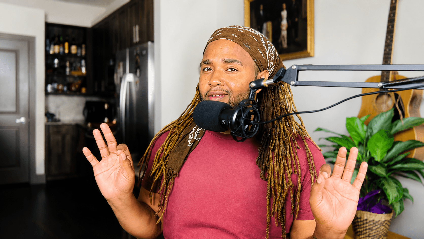 Woman with long hair and headscarf speaking into a microphone, gesturing with hands, in a room with kitchen background.