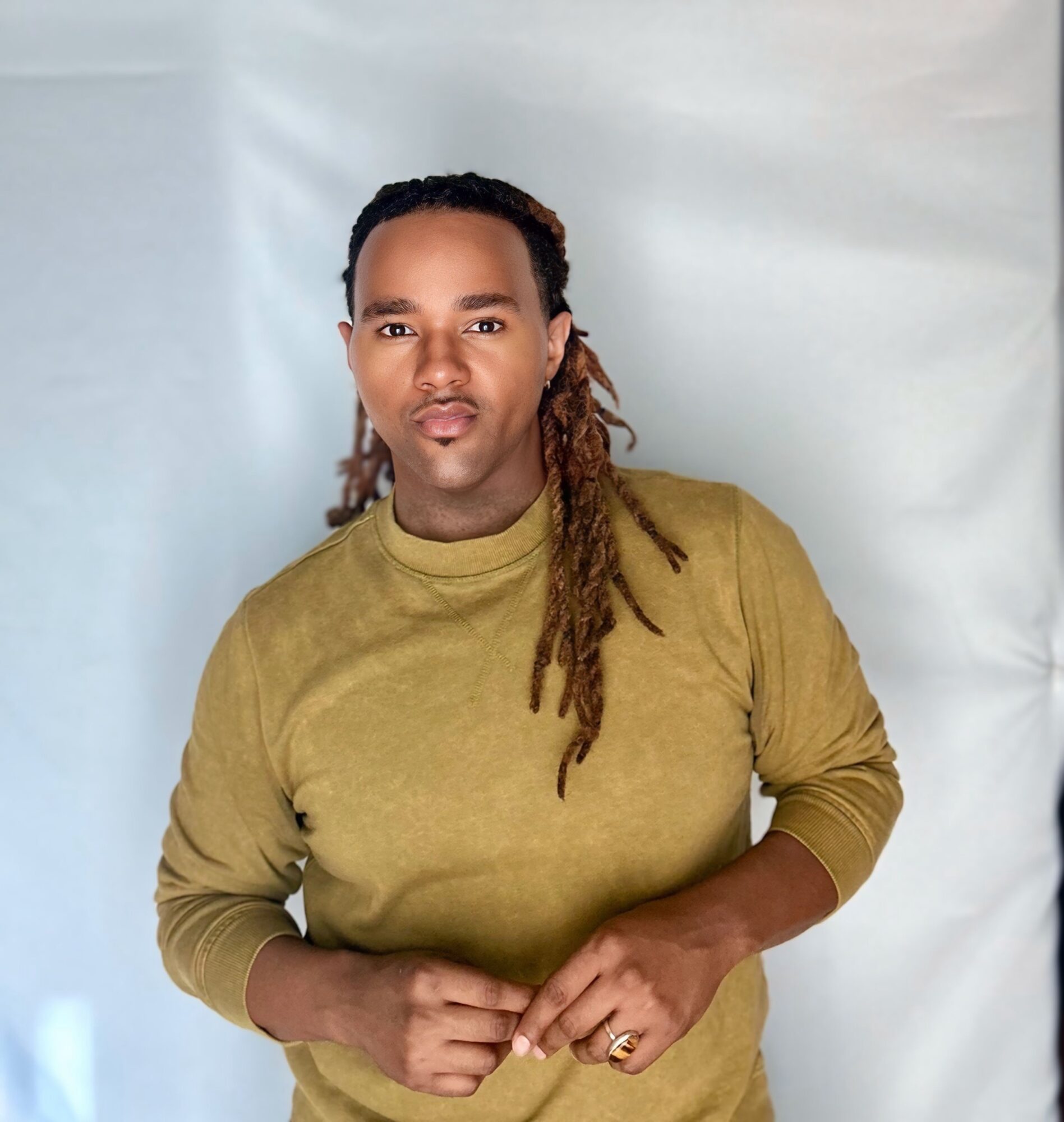 Person with long dreadlocks wearing a mustard-colored top, standing against a light background.