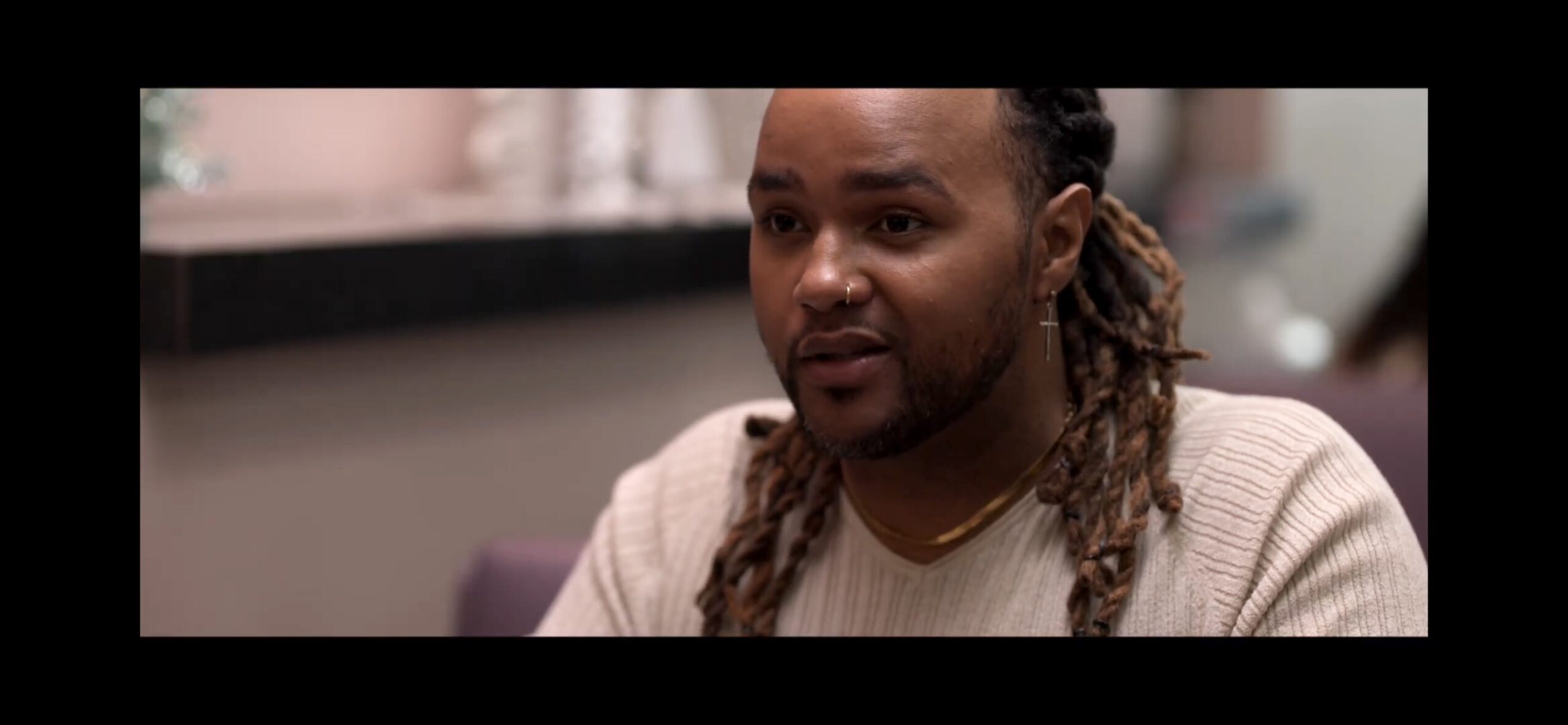 Man with long dreadlocks and a beard sits indoors, wearing a light-colored sweater, looking to the side.