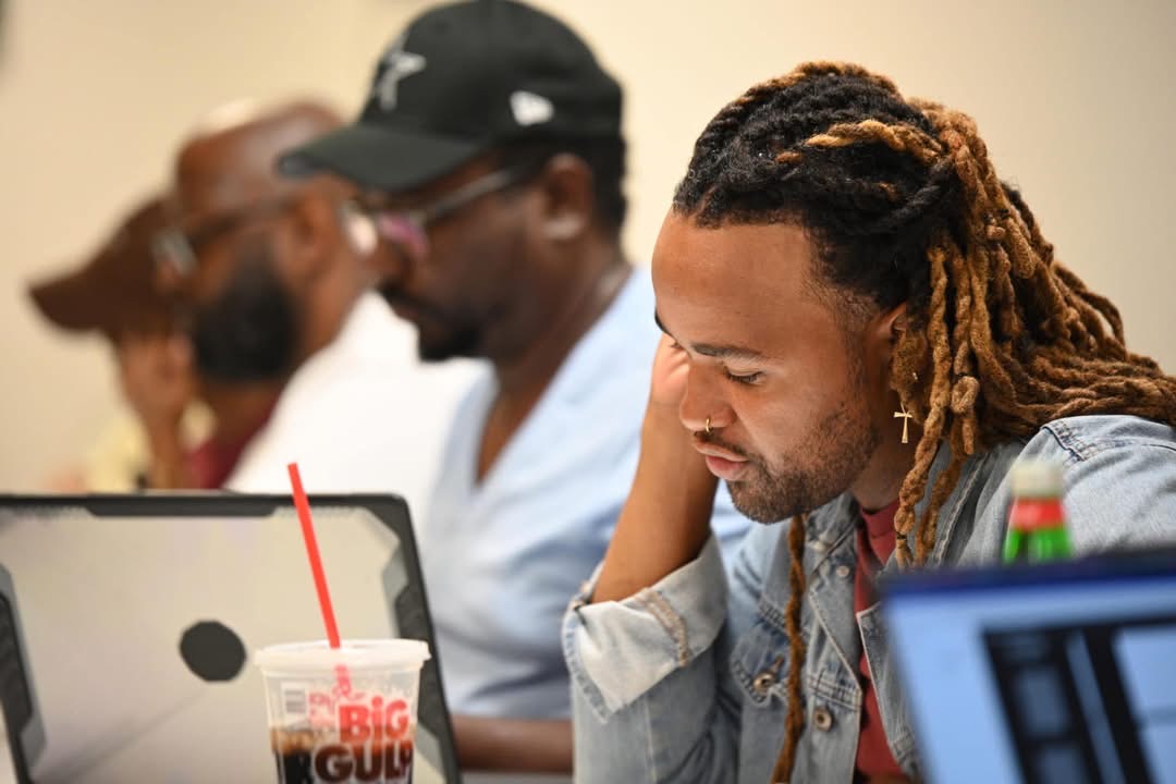 Three people sitting at a table, focused on a laptop, with a drink in the foreground, in a casual setting.