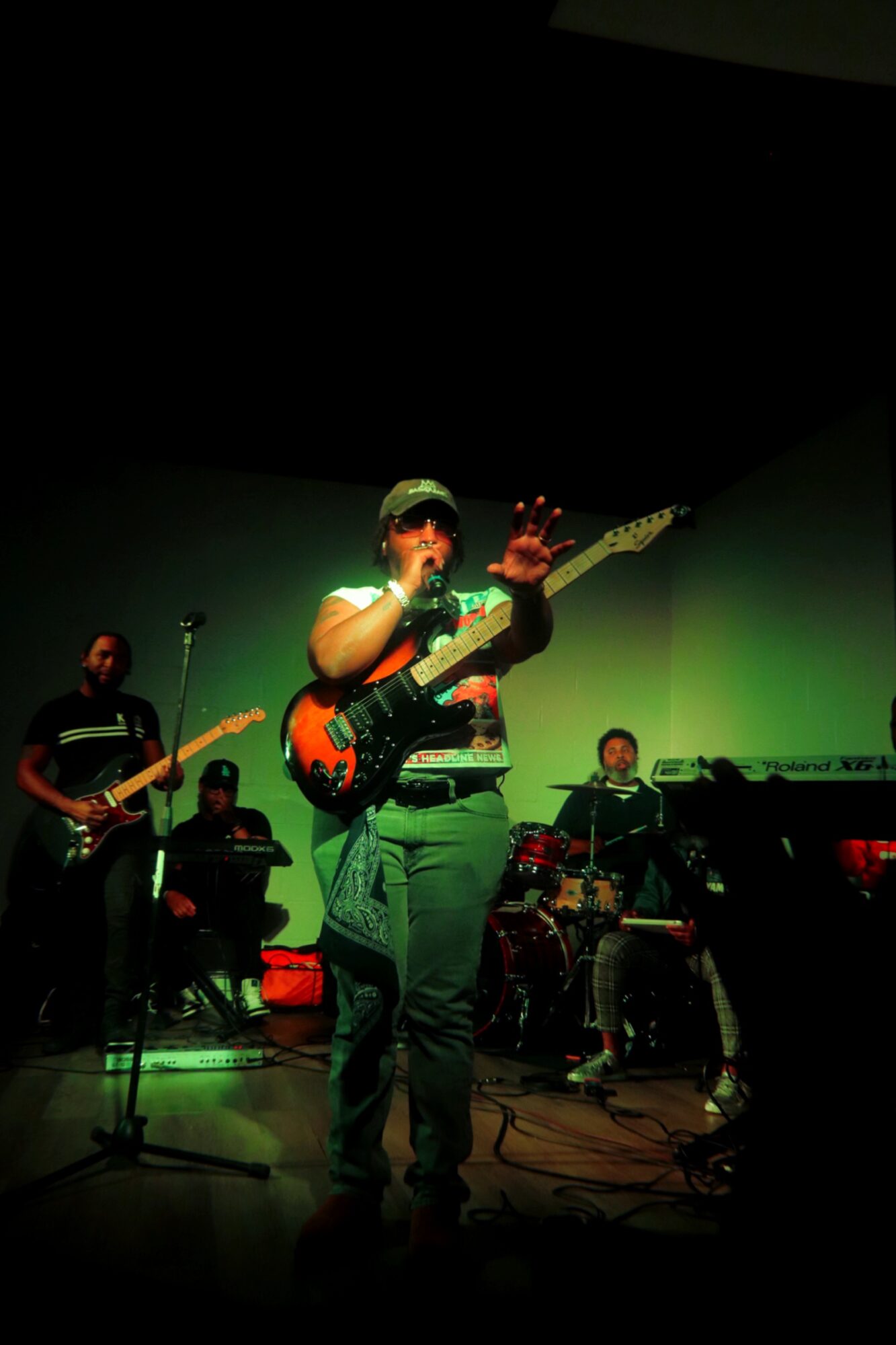 Person playing guitar on stage with band, green lighting, dark background, microphone stand visible.