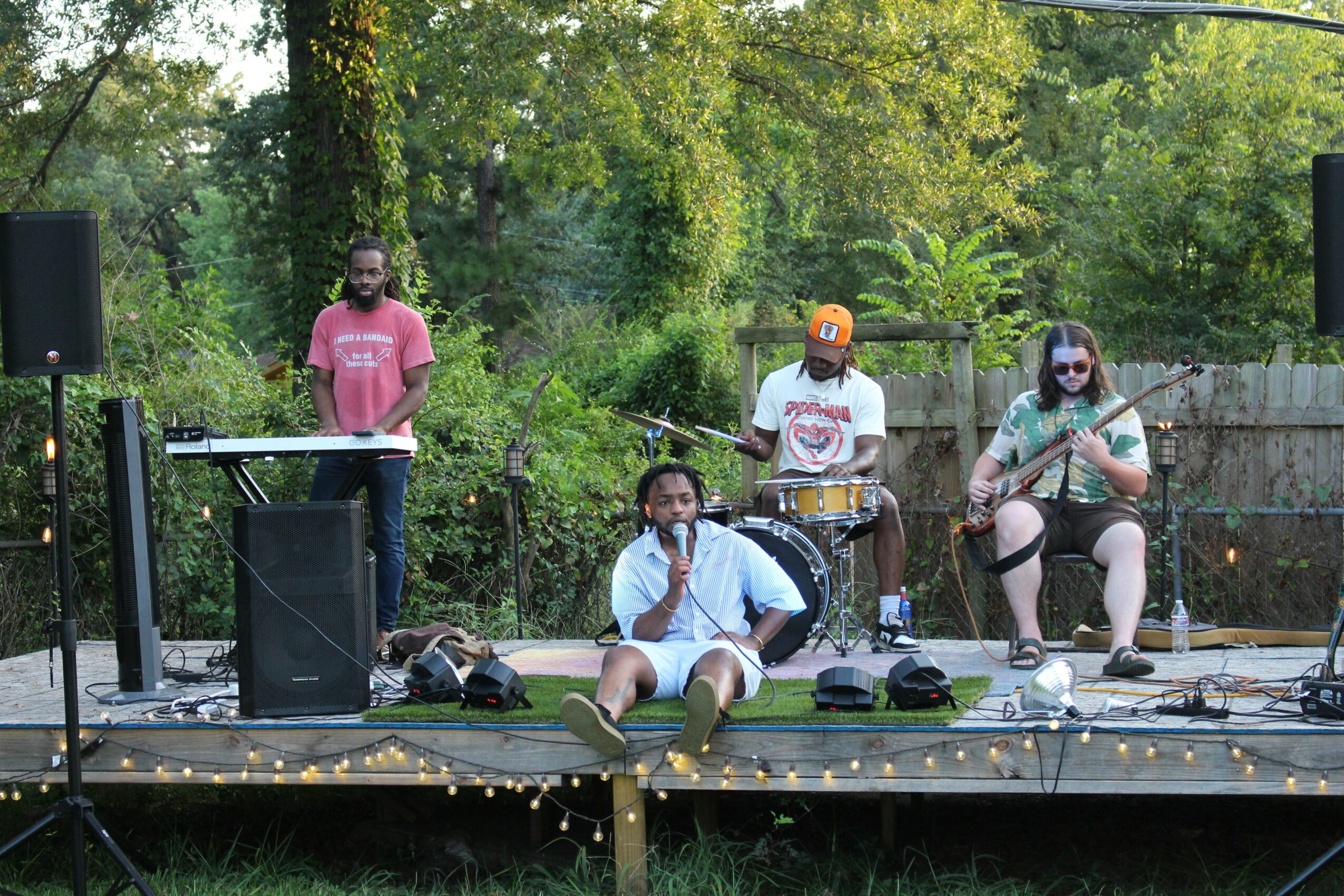Four people performing music on an outdoor stage with trees in the background.