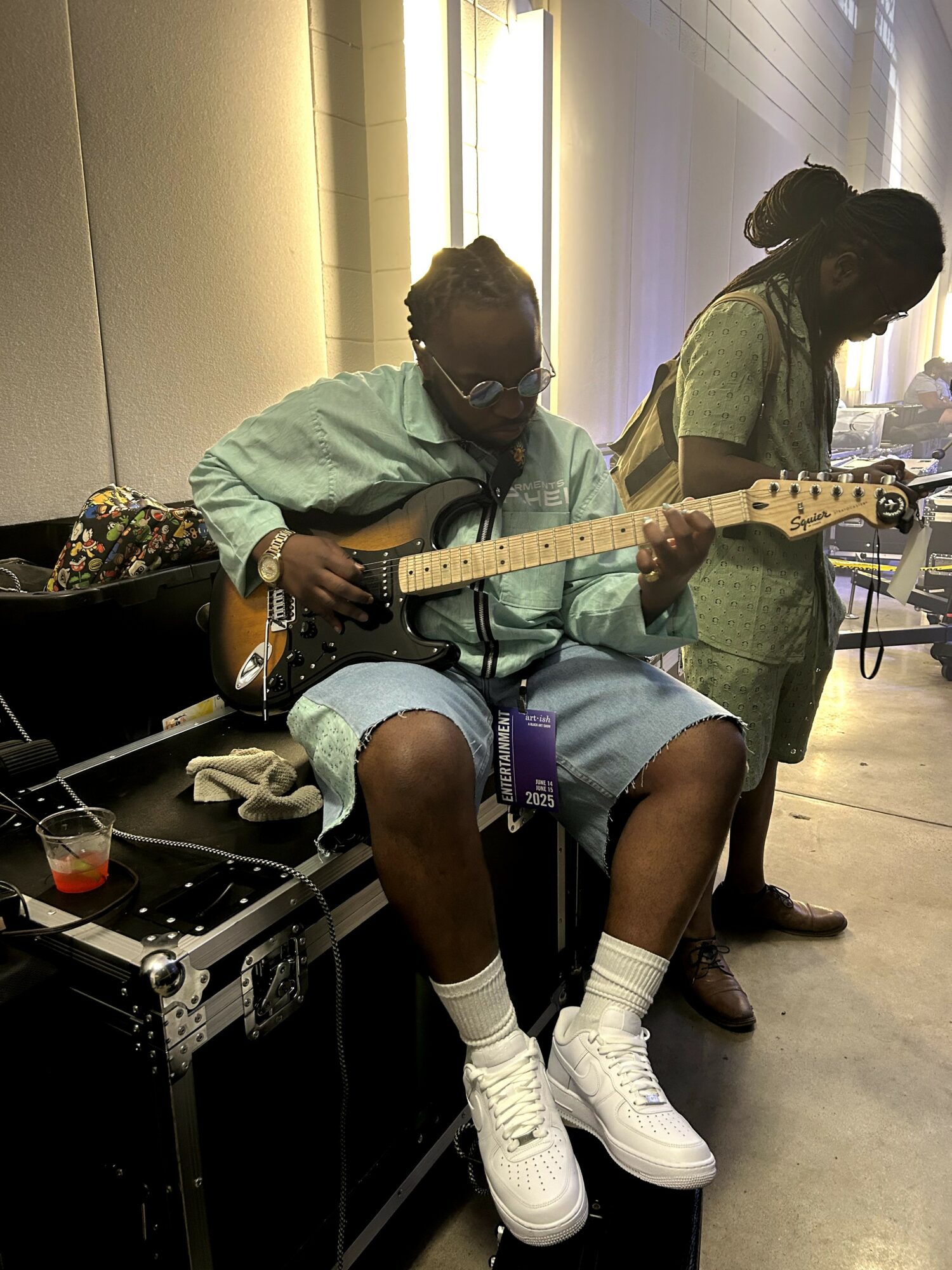 Person sitting on a case playing an electric guitar, another person standing nearby, both in a hallway.