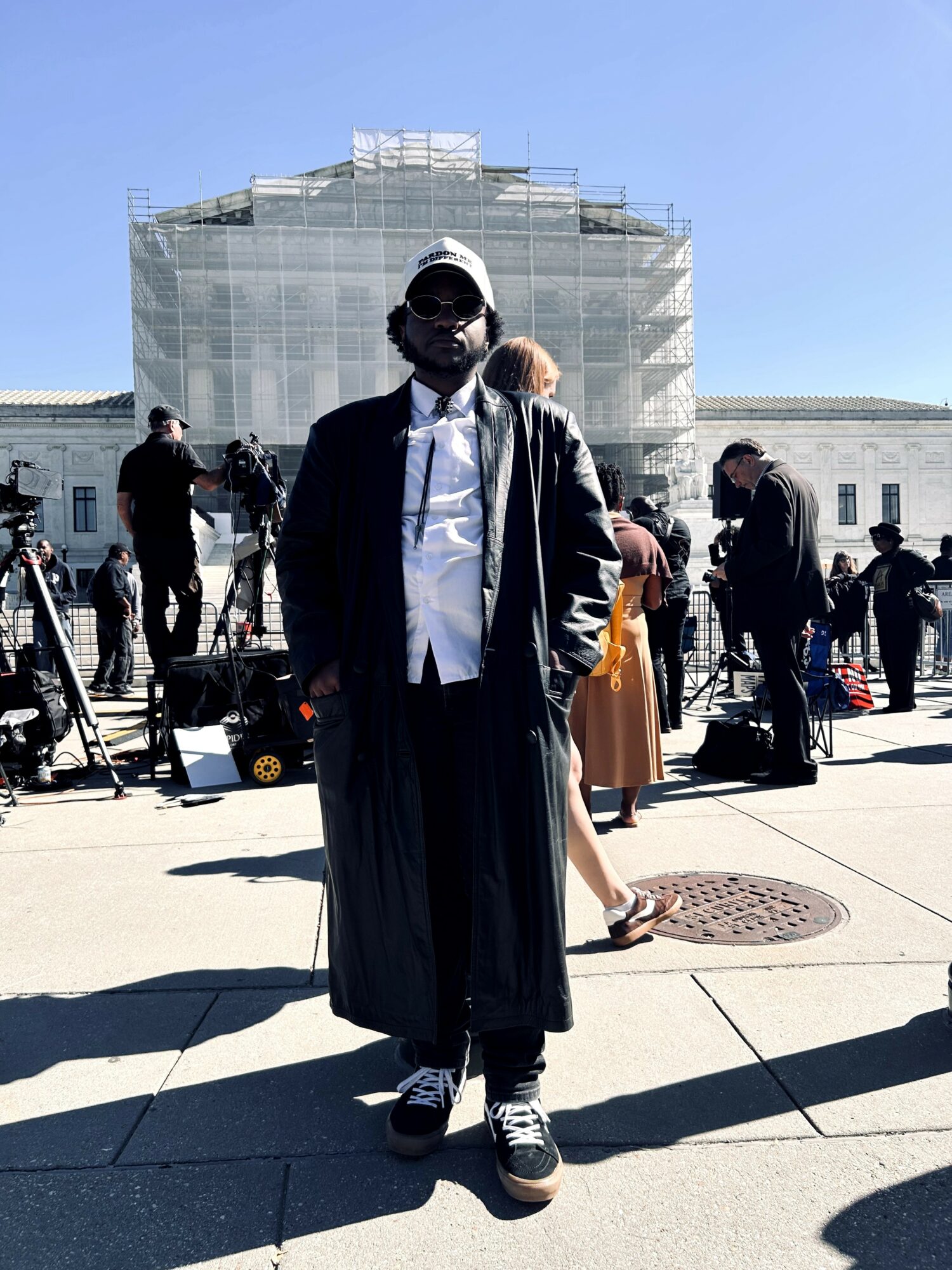 Person standing outdoors in front of a large building, wearing a long coat, sunglasses, and a cap, with people and equipment around.