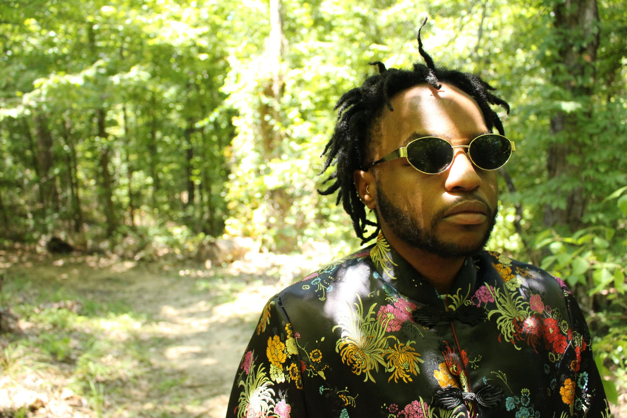 Man with sunglasses and dreadlocks standing outdoors in a forested area, wearing a floral shirt.