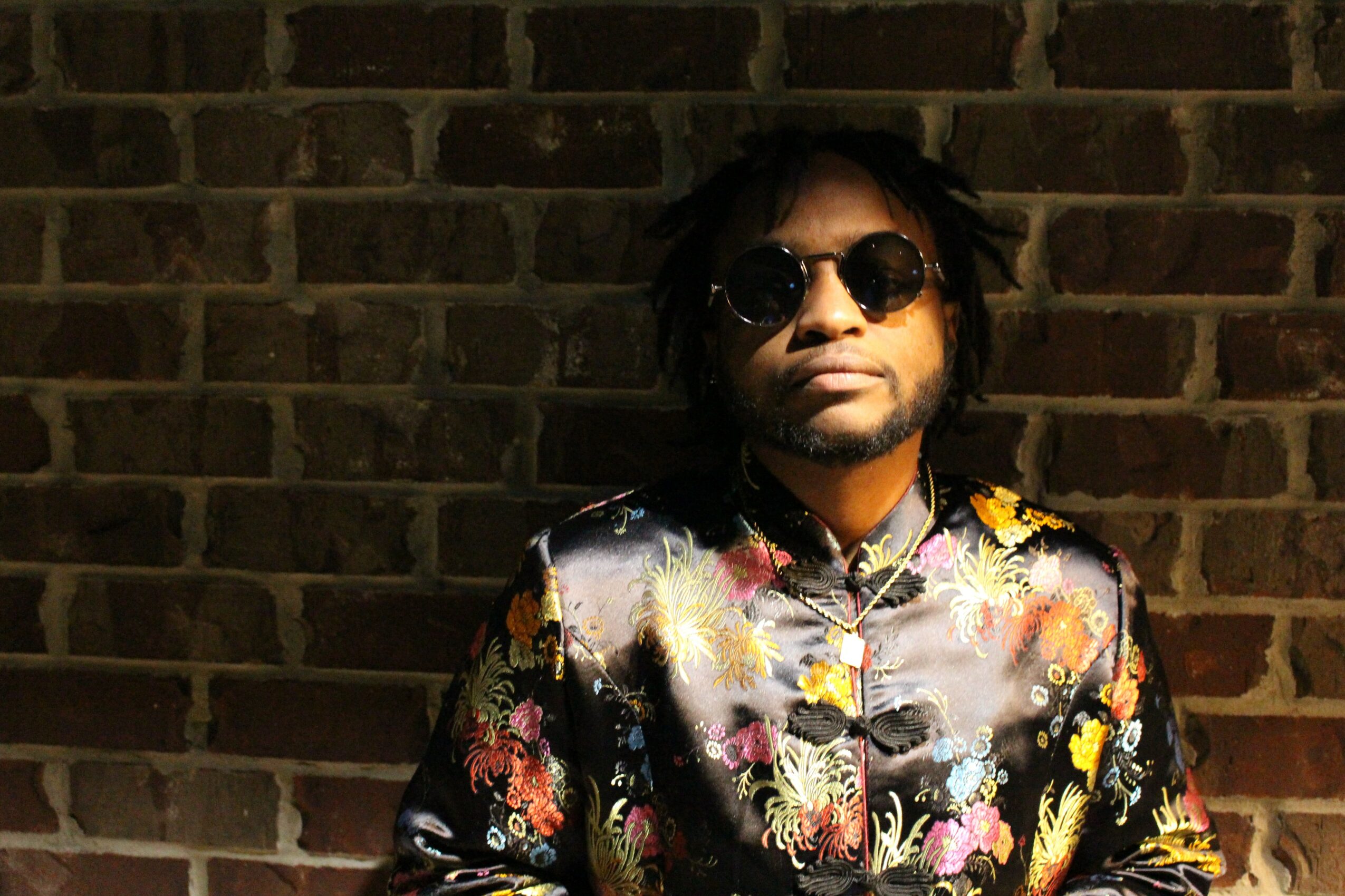 Person with sunglasses and curly hair wearing a colorful patterned shirt against a brick wall.