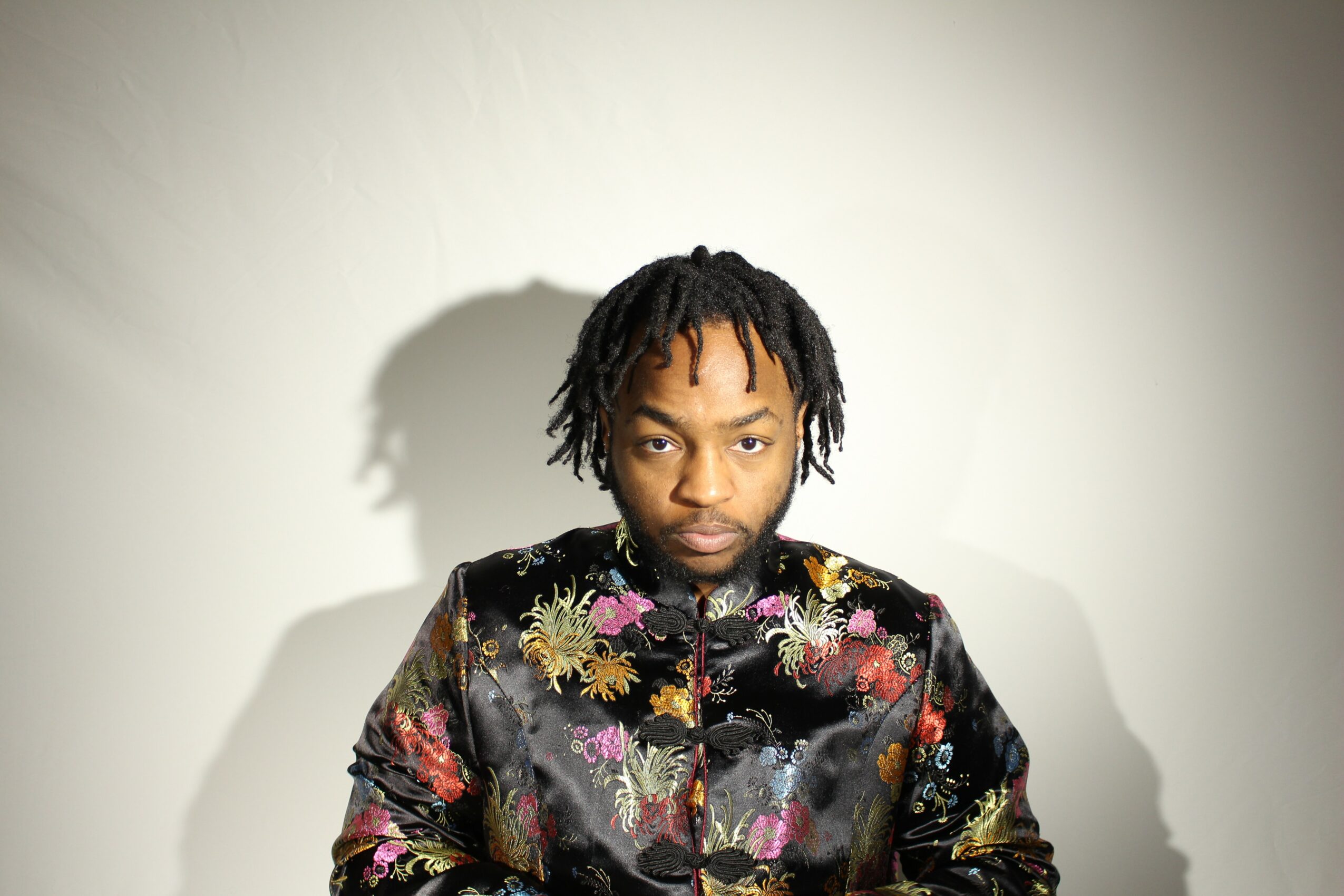 Young man with dreadlocks wearing a colorful floral jacket, standing against a plain background with shadow cast behind him.