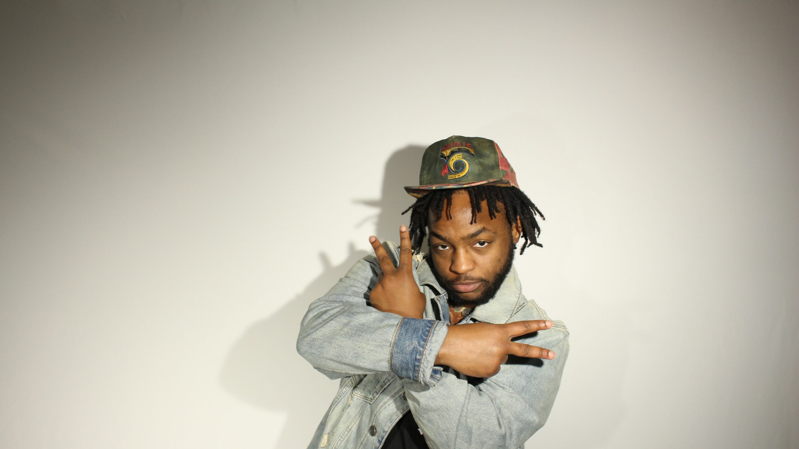 Person with dreadlocks wearing a cap and denim jacket, crossing arms, against a plain background.