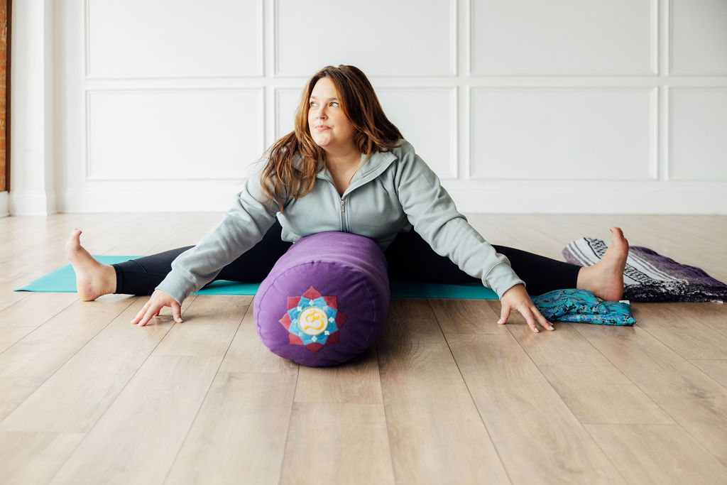 Woman sitting on floor with legs spread, using a purple foam roller, in a bright room with white paneled walls.
