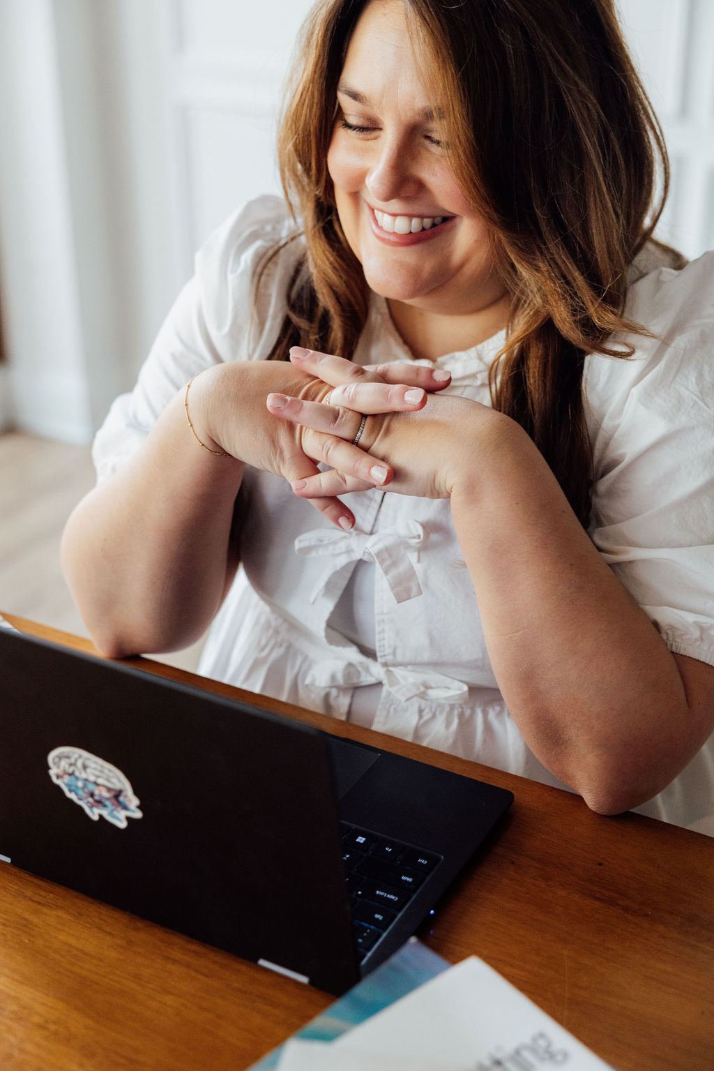 Woman smiling at a laptop, hands clasped, sitting at a wooden table, with a white background.