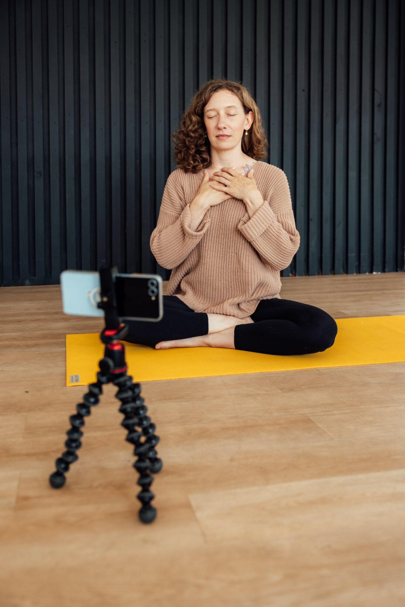Woman practicing yoga on yellow mat, sitting cross-legged with hands on chest, in front of black wall, recording with smartphone on tripod.