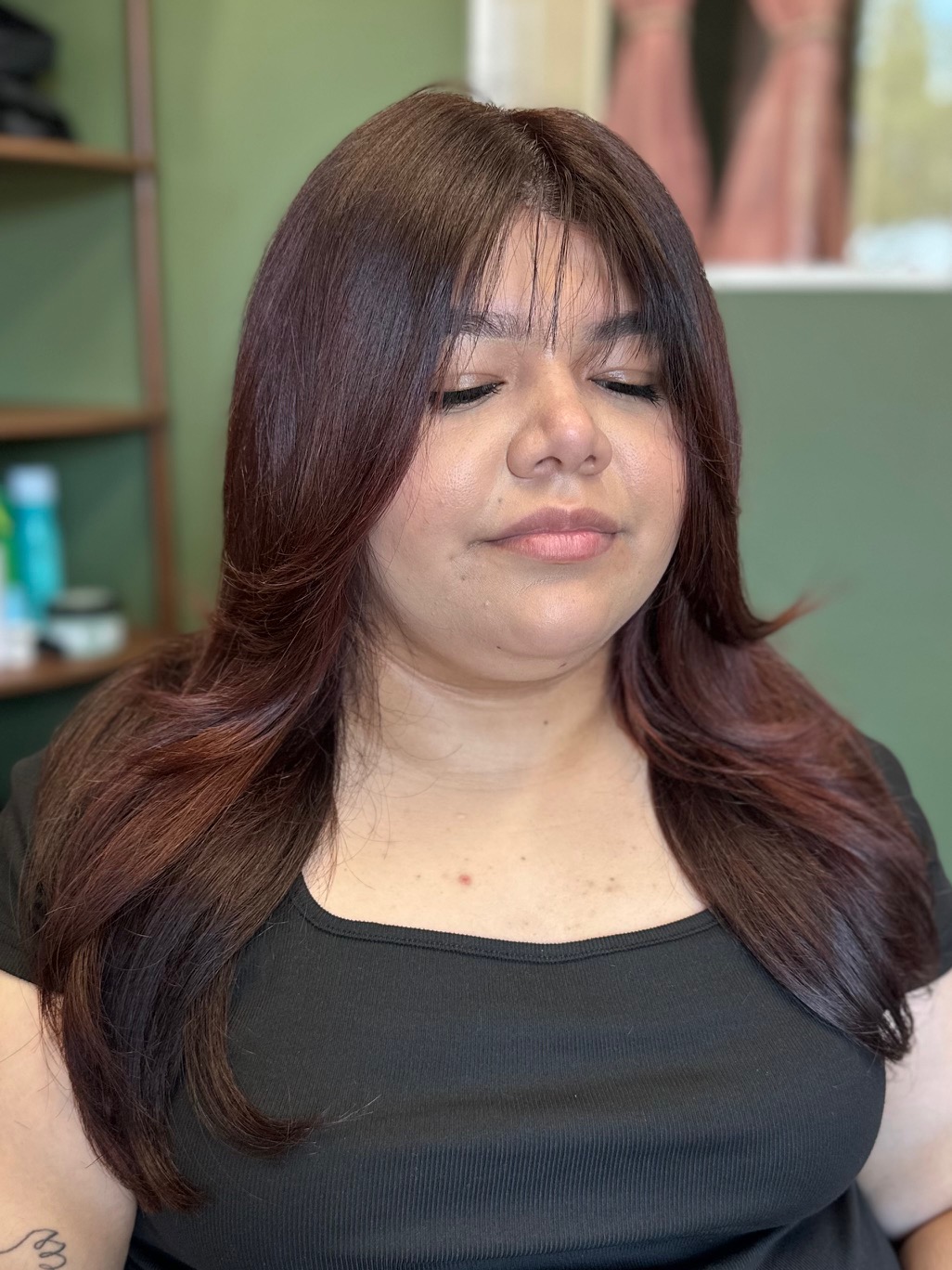 Woman with long, layered brown hair and closed eyes, wearing a black top, in a room with green walls and shelves.