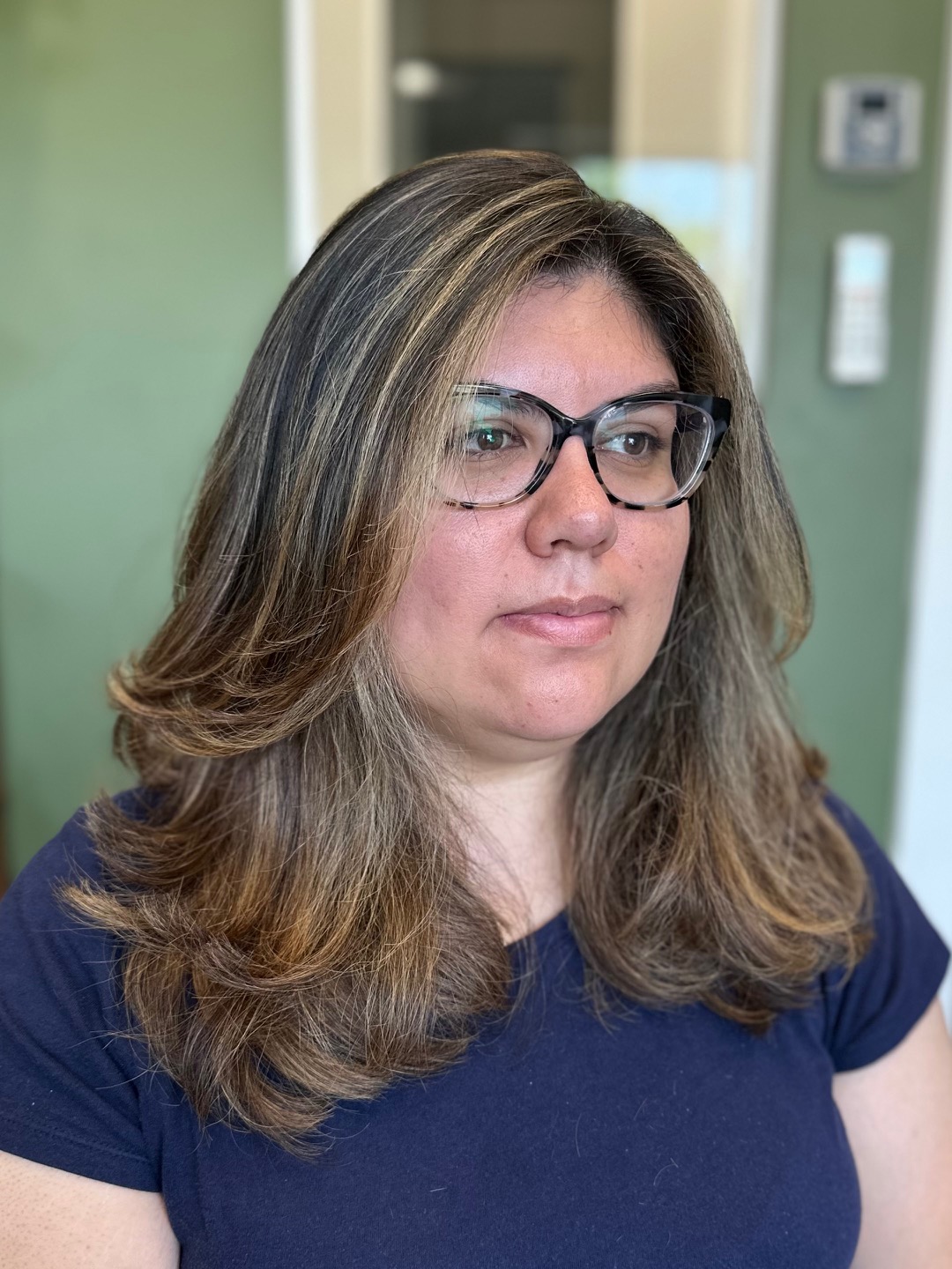 Woman with shoulder-length wavy hair, glasses, and a navy shirt, indoors with green walls and a door in background.