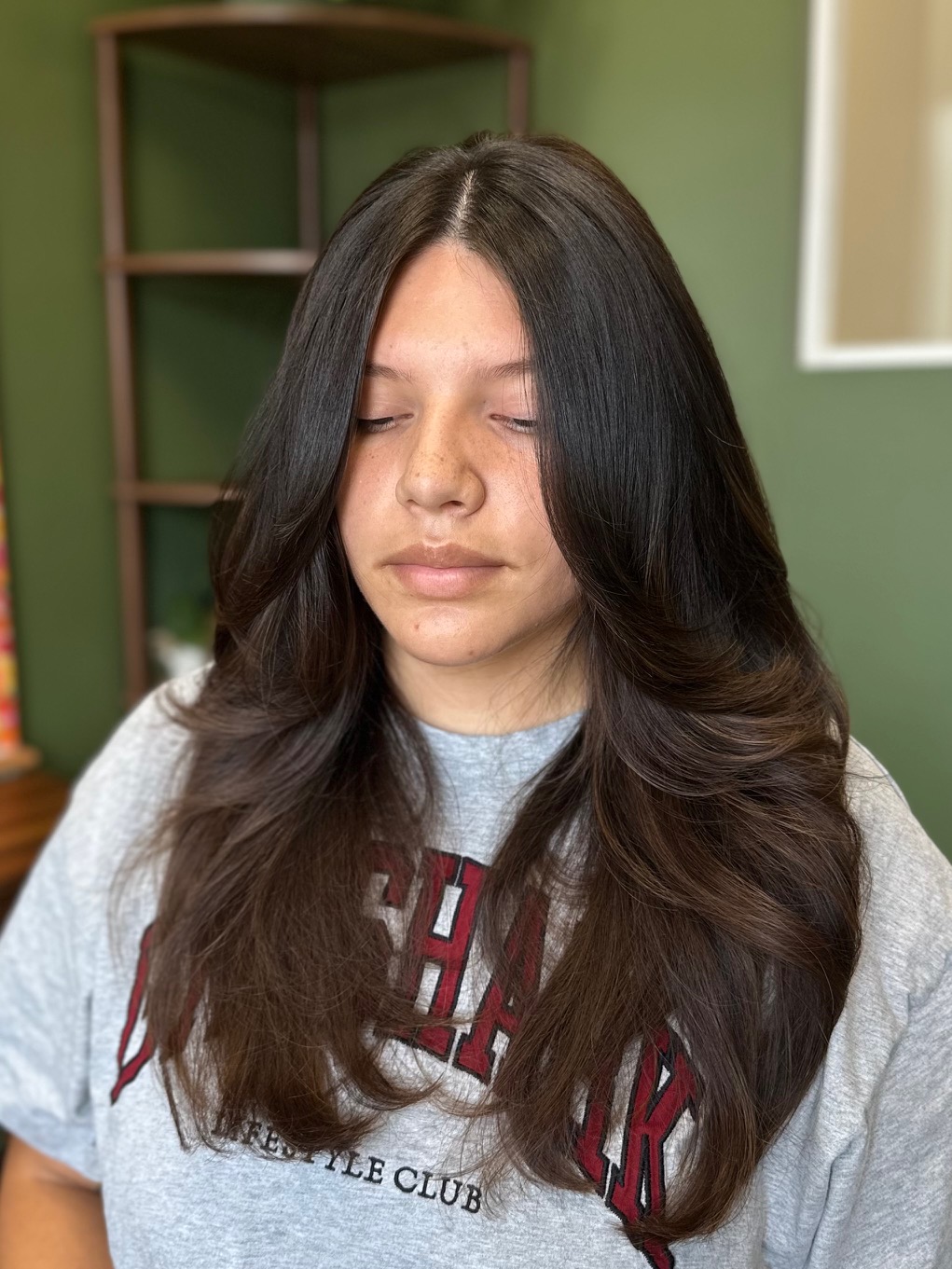 Young woman with long, dark hair and closed eyes, wearing a gray T-shirt, in a room with green walls.