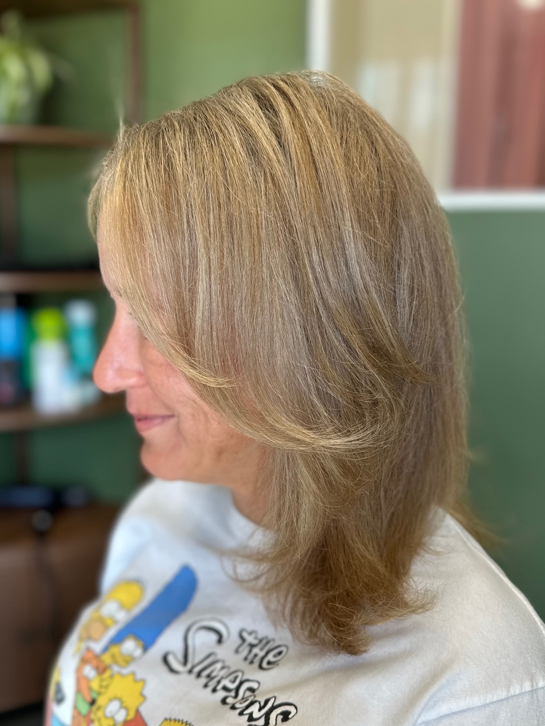 Side view of a woman with shoulder-length blonde hair in a salon, wearing a white shirt with cartoon characters.
