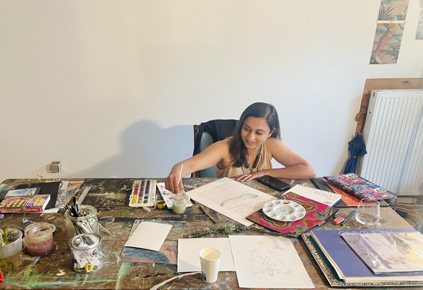 Girl sitting at a cluttered table with art supplies, papers, and books, reaching for something on the table.