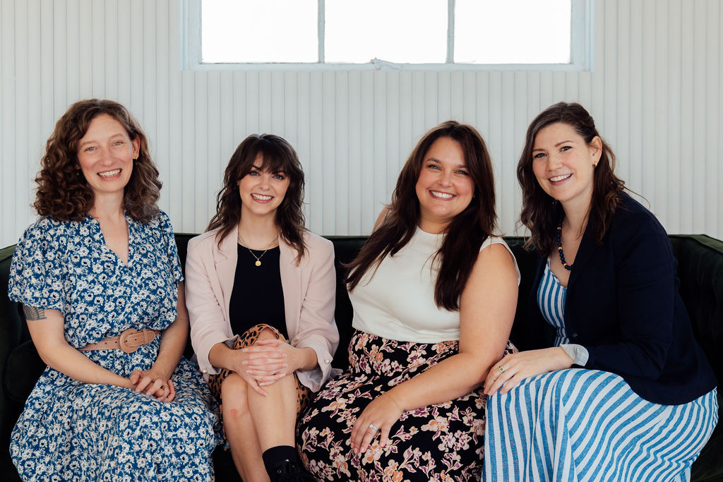 Four women sitting together indoors, smiling, with a window behind them, dressed in casual and semi-formal clothing.