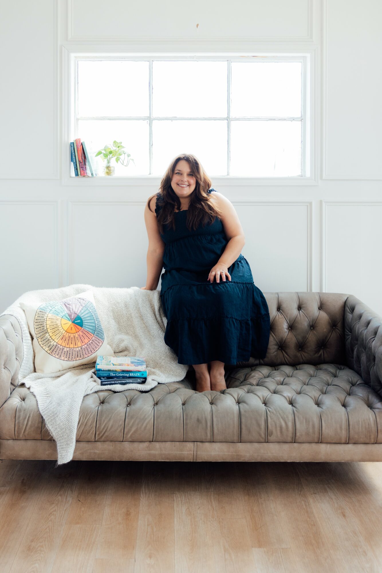 Woman sitting on a tufted sofa in a bright room with a large window behind her, books and a plant on the windowsill.