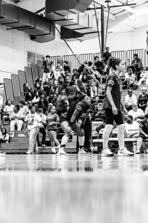 Indoor sports event with players, coaches, and spectators seated in bleachers, some standing on the court, black and white photo.
