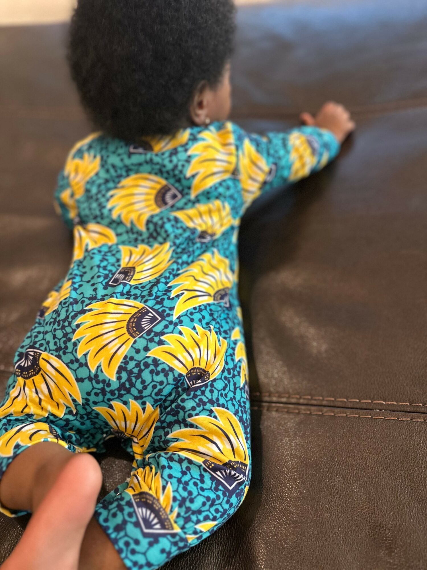 Child lying face down on a brown surface, wearing a colorful dress with yellow and blue patterns.