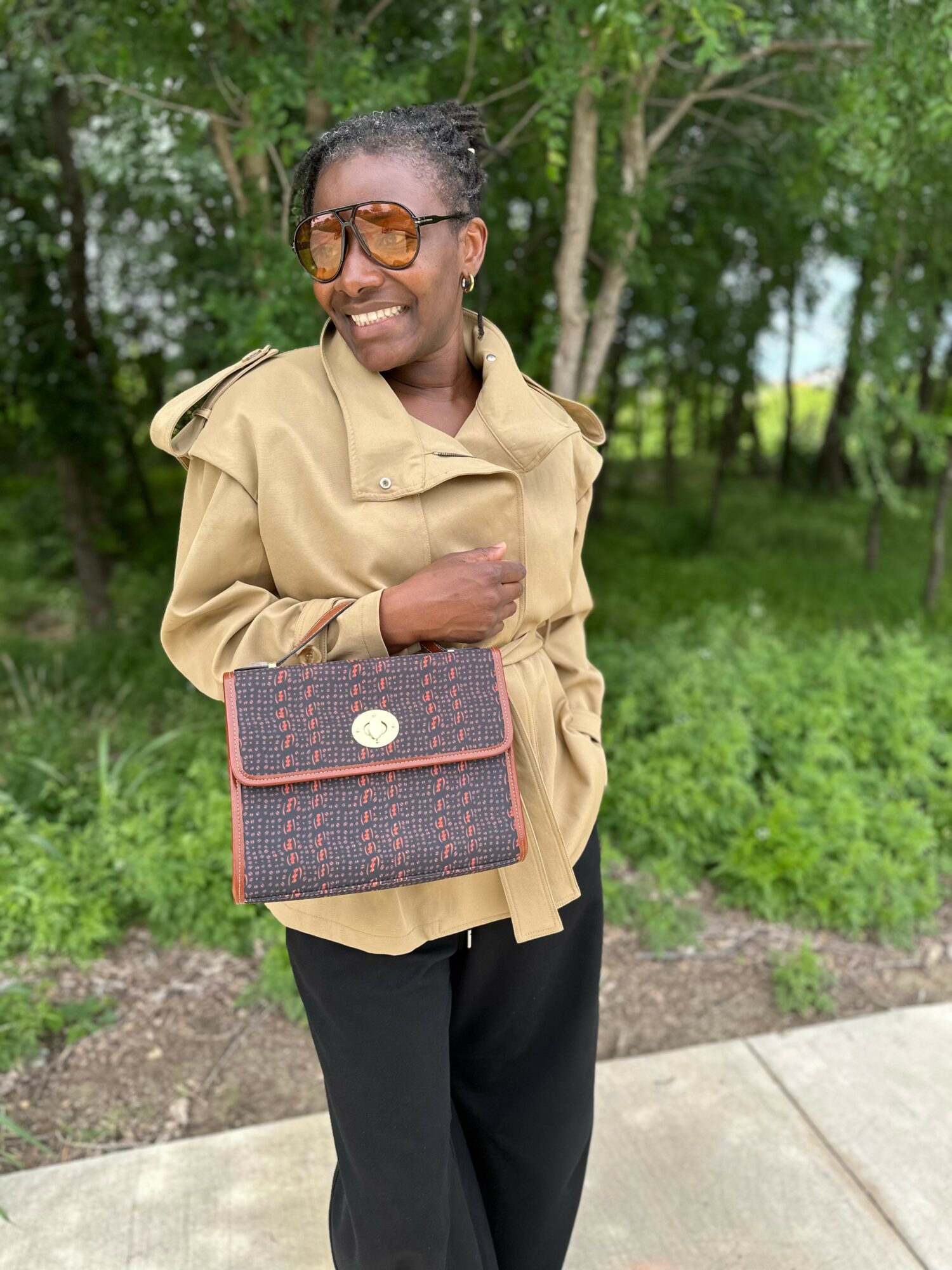 Woman outdoors smiling, wearing sunglasses, beige jacket, black pants, holding a patterned handbag, trees in background.