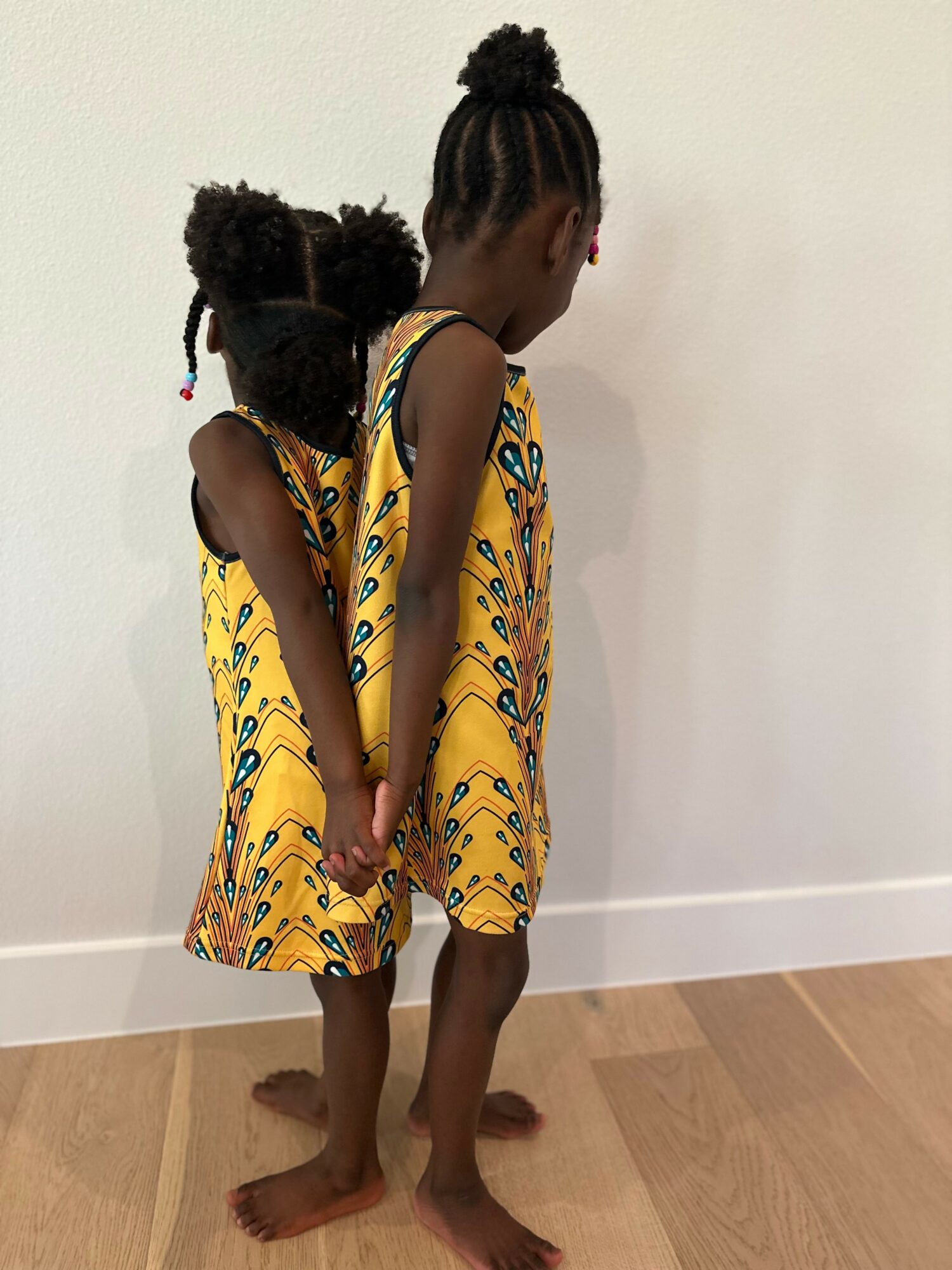 Two children in matching yellow patterned dresses stand side by side, facing away from the camera, holding hands.
