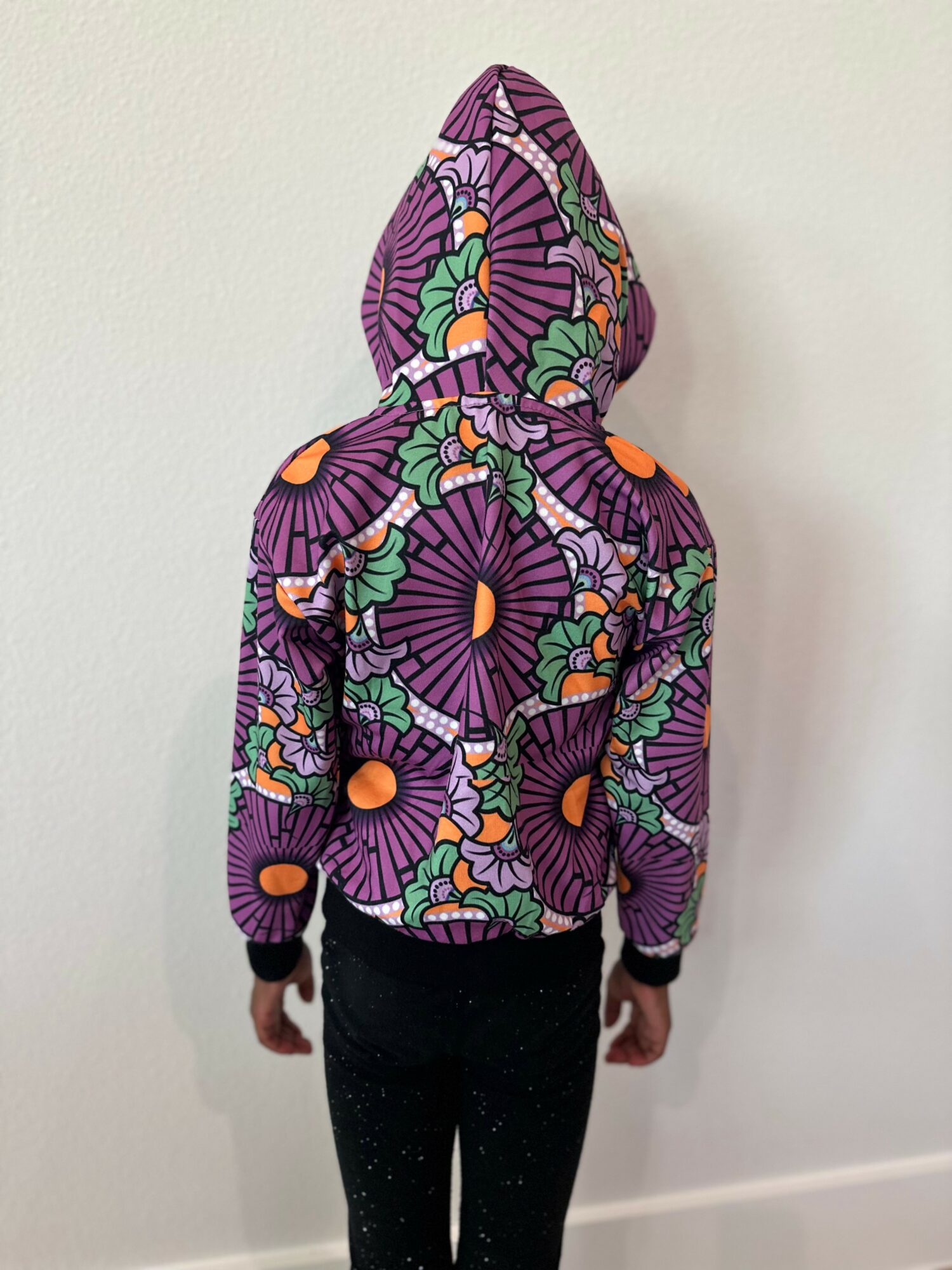 Person wearing a colorful, patterned hoodie with a floral design, facing a plain white wall.