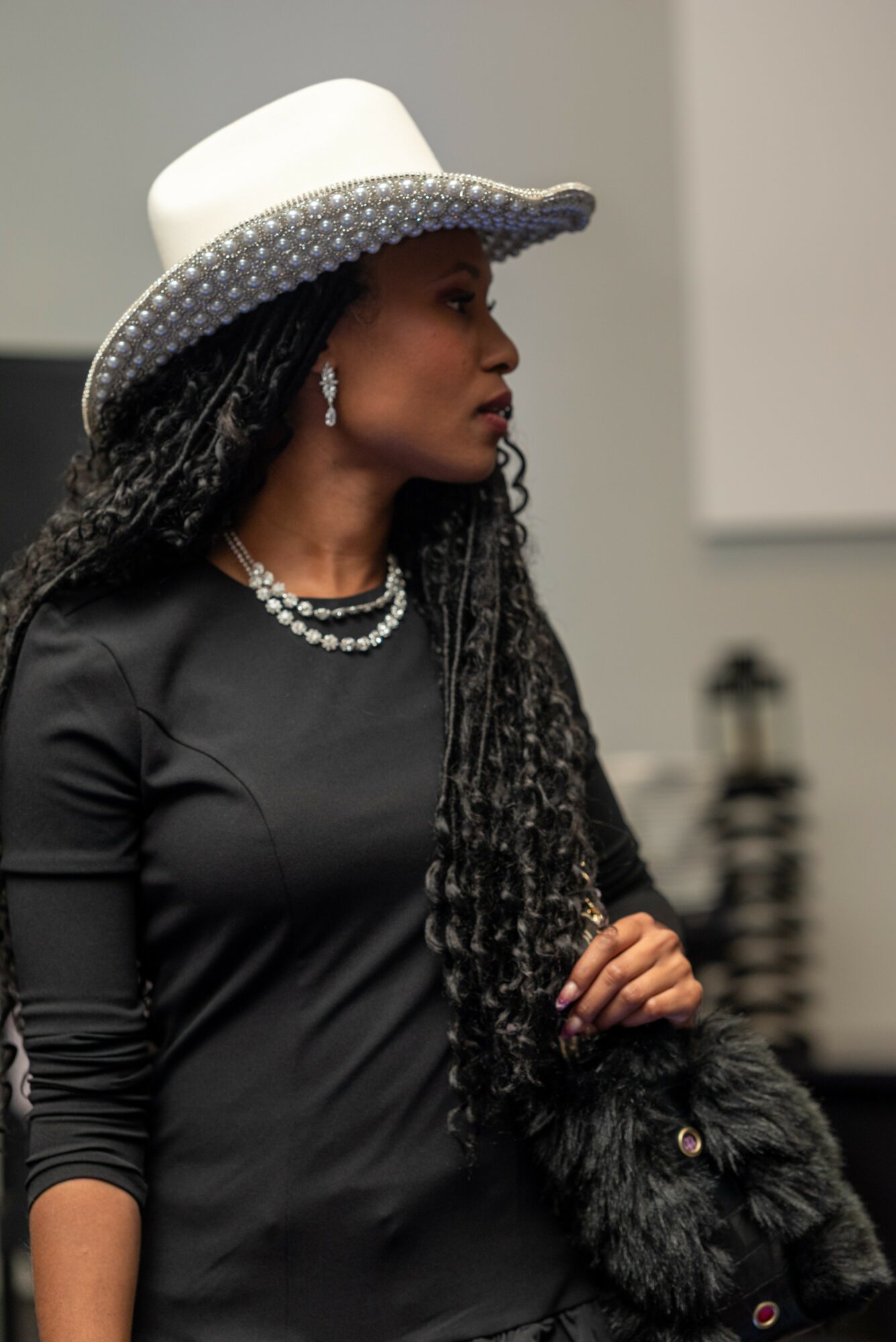 Woman wearing a white hat with a silver band, black dress, pearl necklace, and earrings, holding a black furry object.