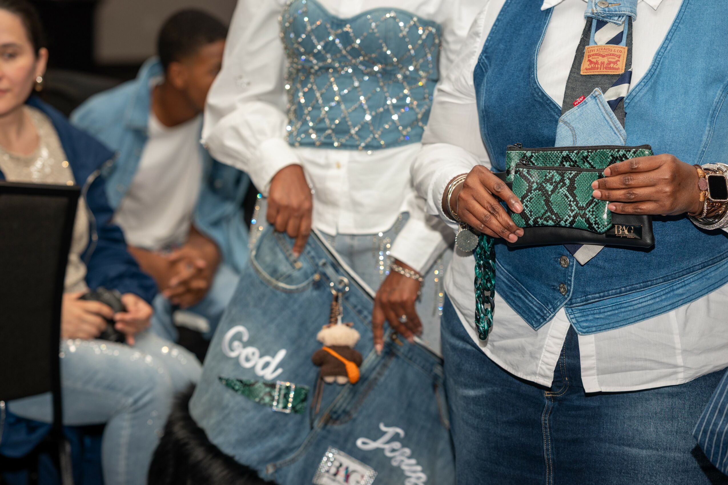 People gathered at an event, with focus on a person holding a green patterned clutch and wearing a blue vest.