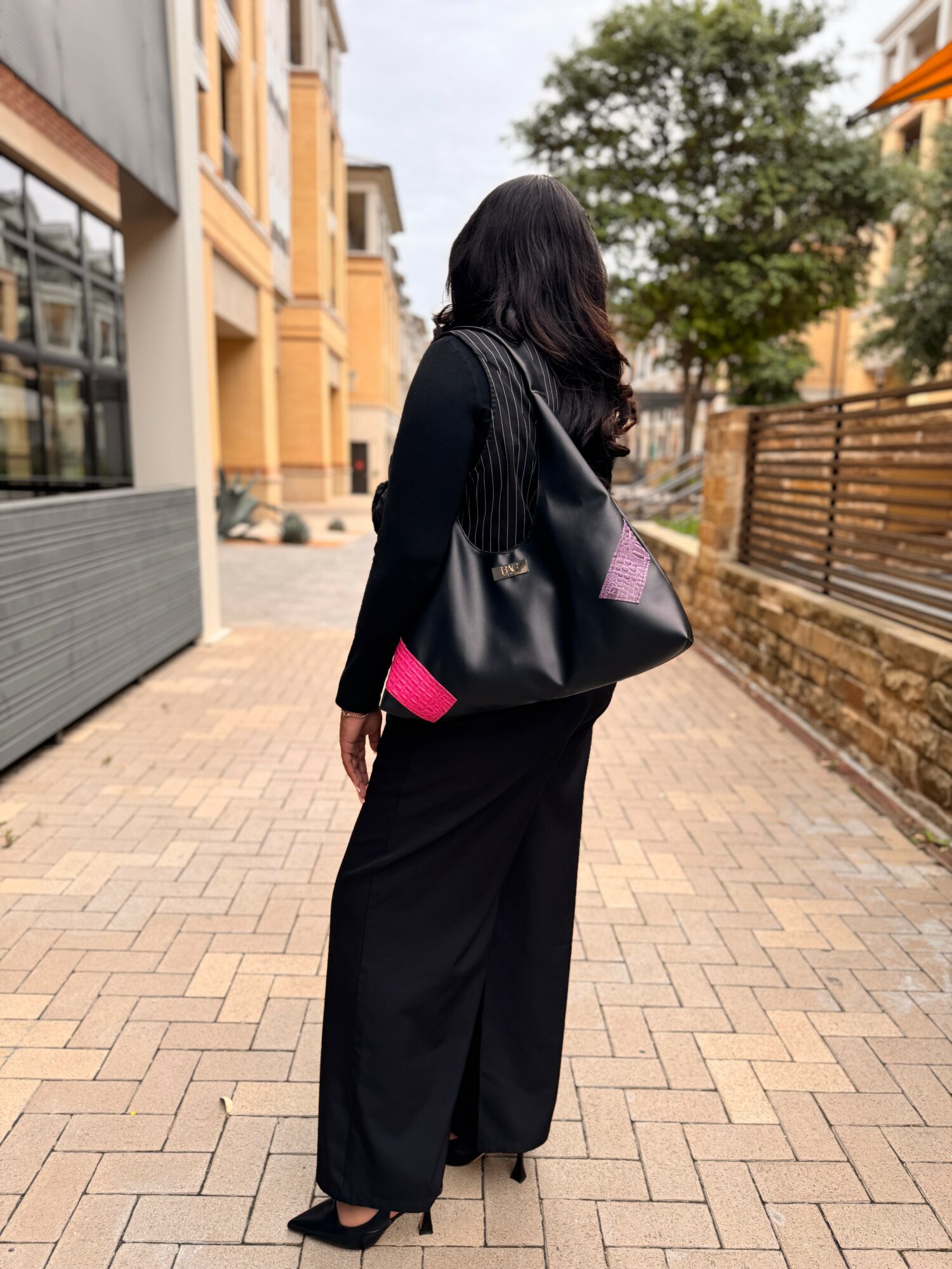 Woman with long dark hair walking on a brick sidewalk, wearing a black dress and carrying a black bag with pink accents, in an urban setting with buildings and trees.