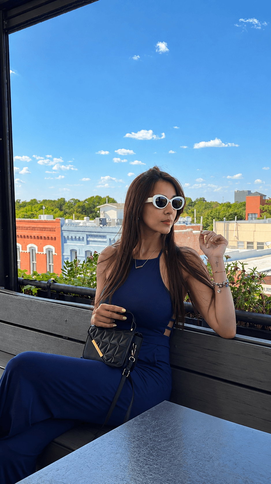 Woman with long hair wearing sunglasses and a blue dress, sitting outdoors with a cityscape and blue sky in background.