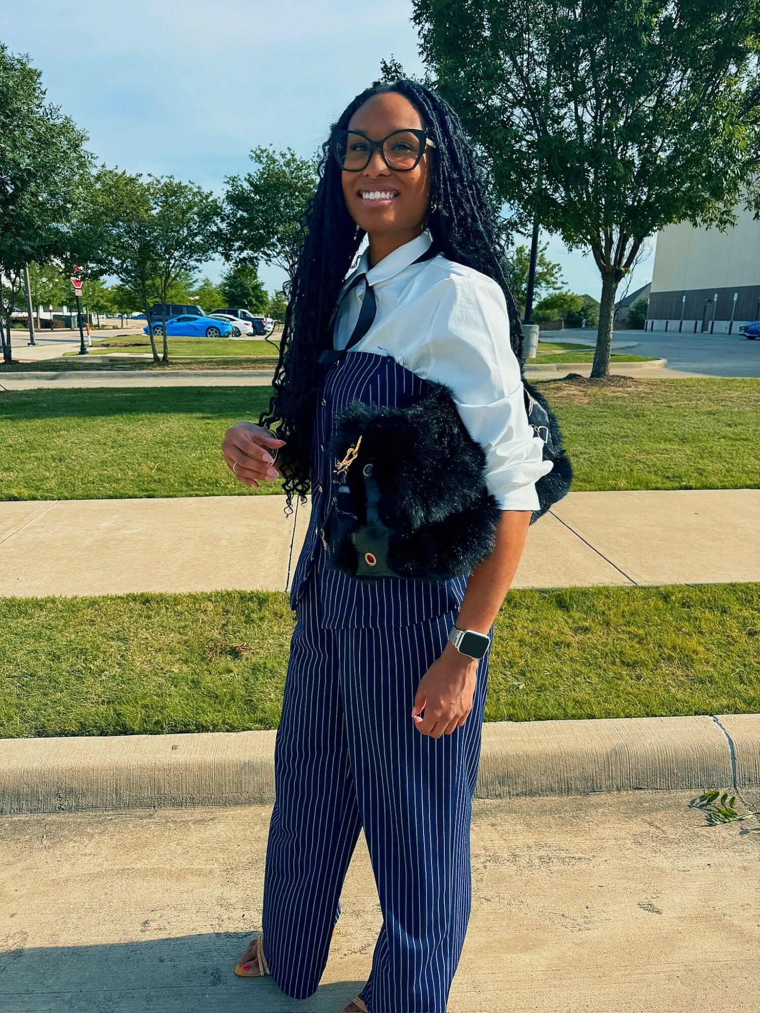 Woman with glasses and braided hair smiling outdoors, wearing a white shirt and striped pants, holding a black purse.